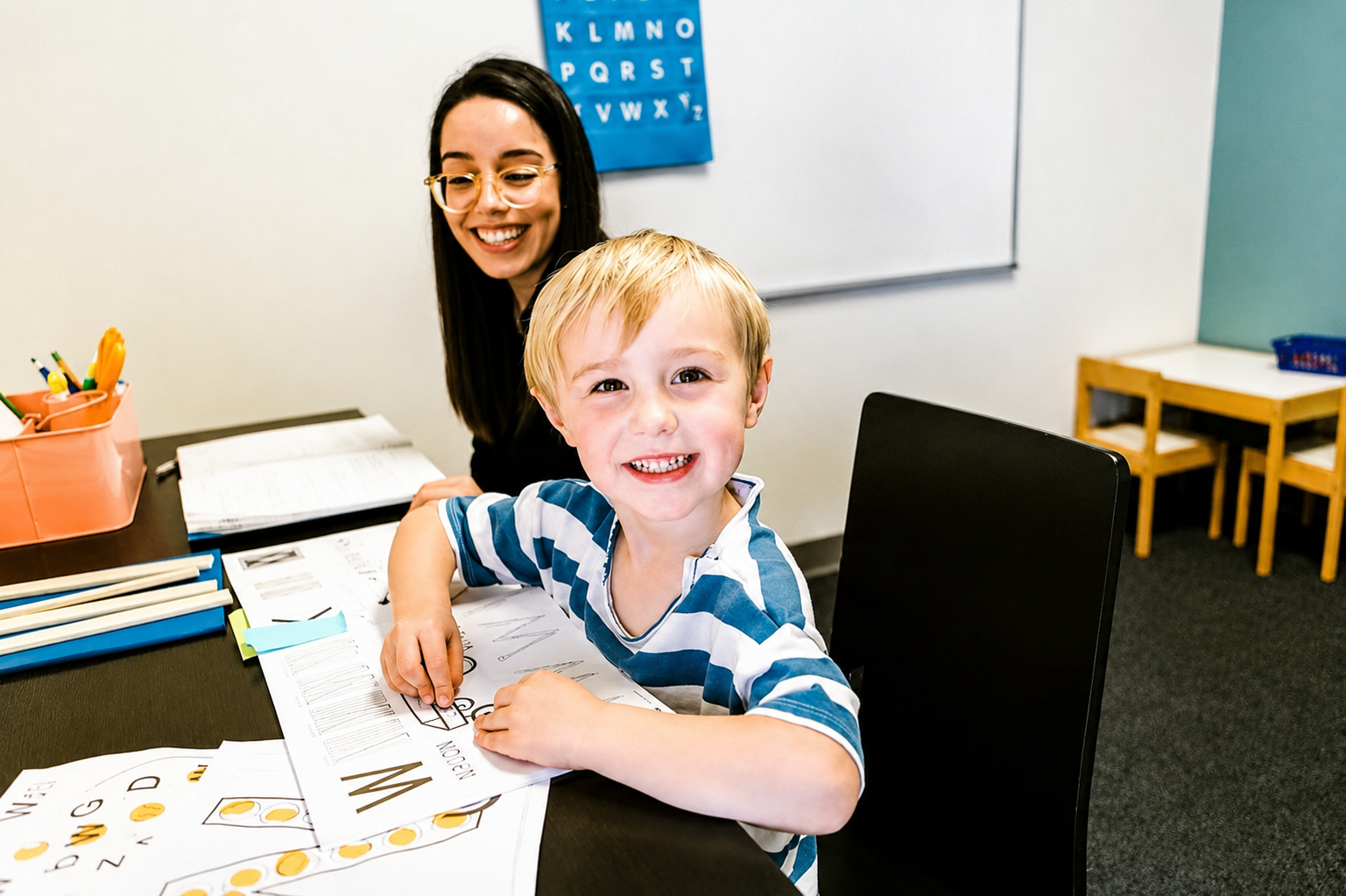 A young boy and a woman, likely a teacher, working on alphabet worksheets in a classroom. The boy is smiling at the camera, wearing a blue and white striped shirt. The woman is smiling and wearing glasses, sitting behind him with a blue alphabet chart on the wall.