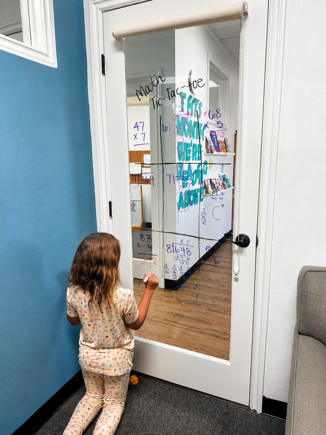 A young girl in pajamas kneeling and writing on a clear glass door with a dry erase marker. The door has colorful math problems and words written on it, including 'Math Tic-Tac-toe' and 'You are here.'
