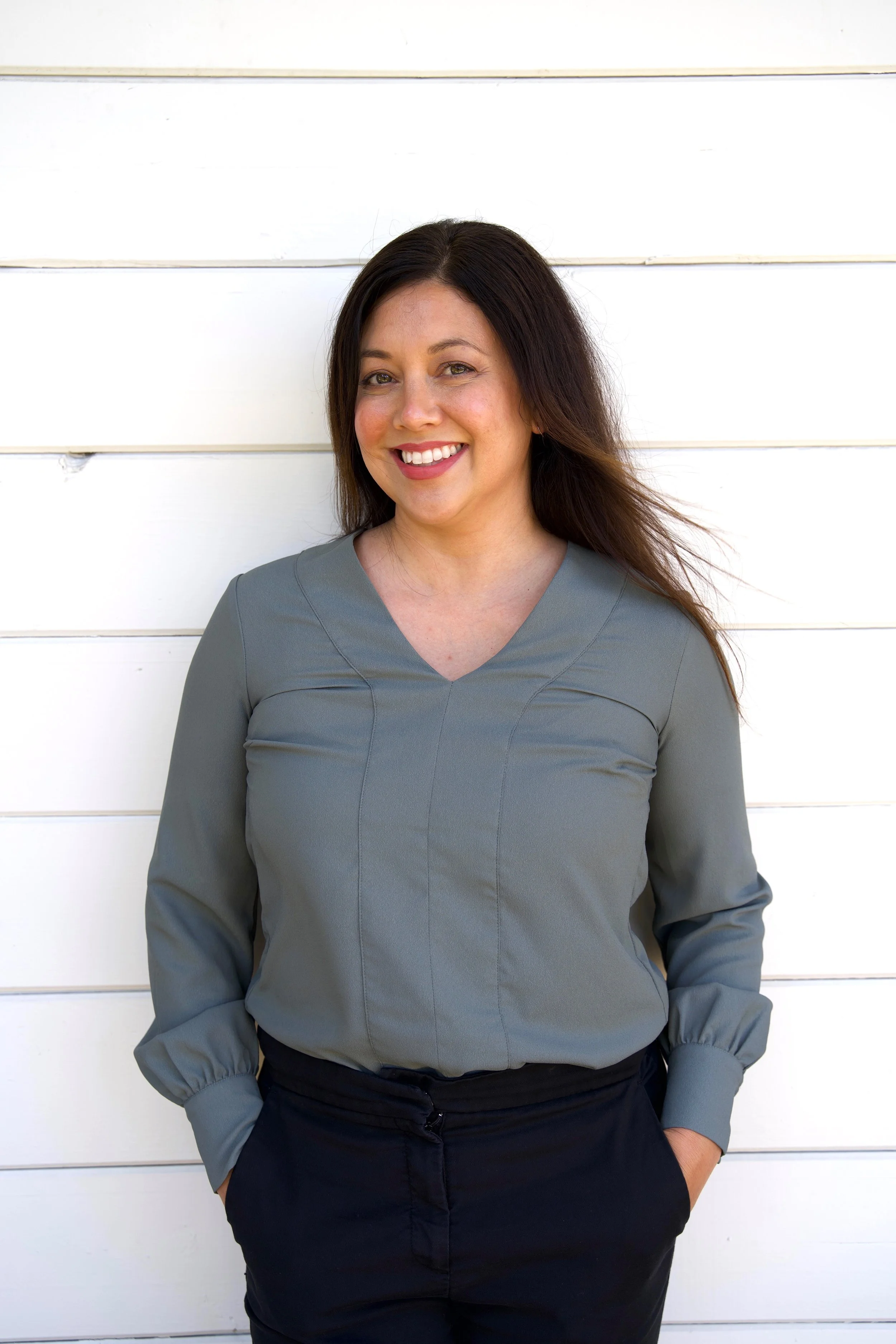 A woman with long dark hair smiling and looking at the camera, standing against a white wooden wall, wearing a gray long-sleeve top and black pants.