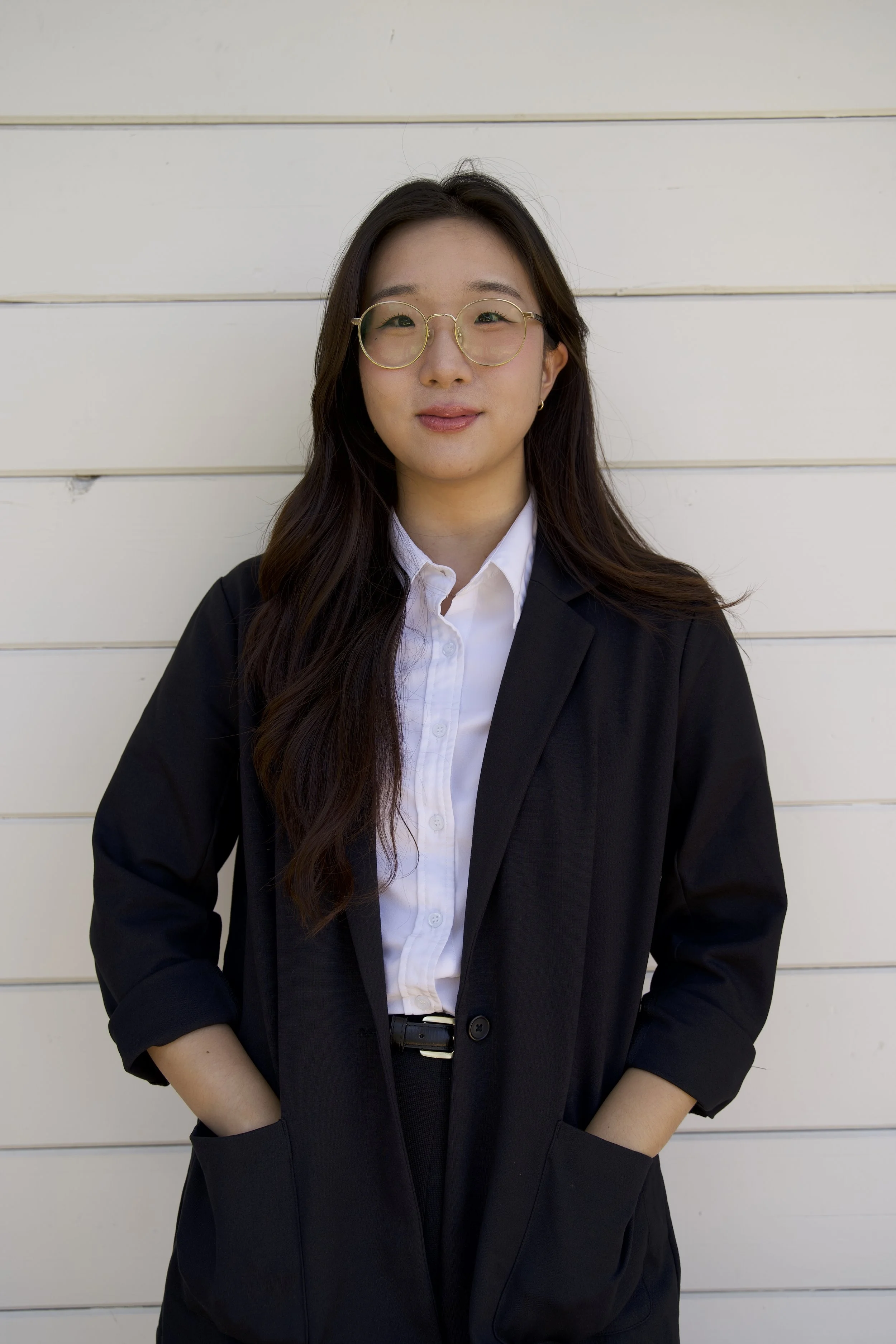 A young woman with long dark hair, wearing round glasses, a white shirt, and a black blazer, standing in front of a white wooden wall.