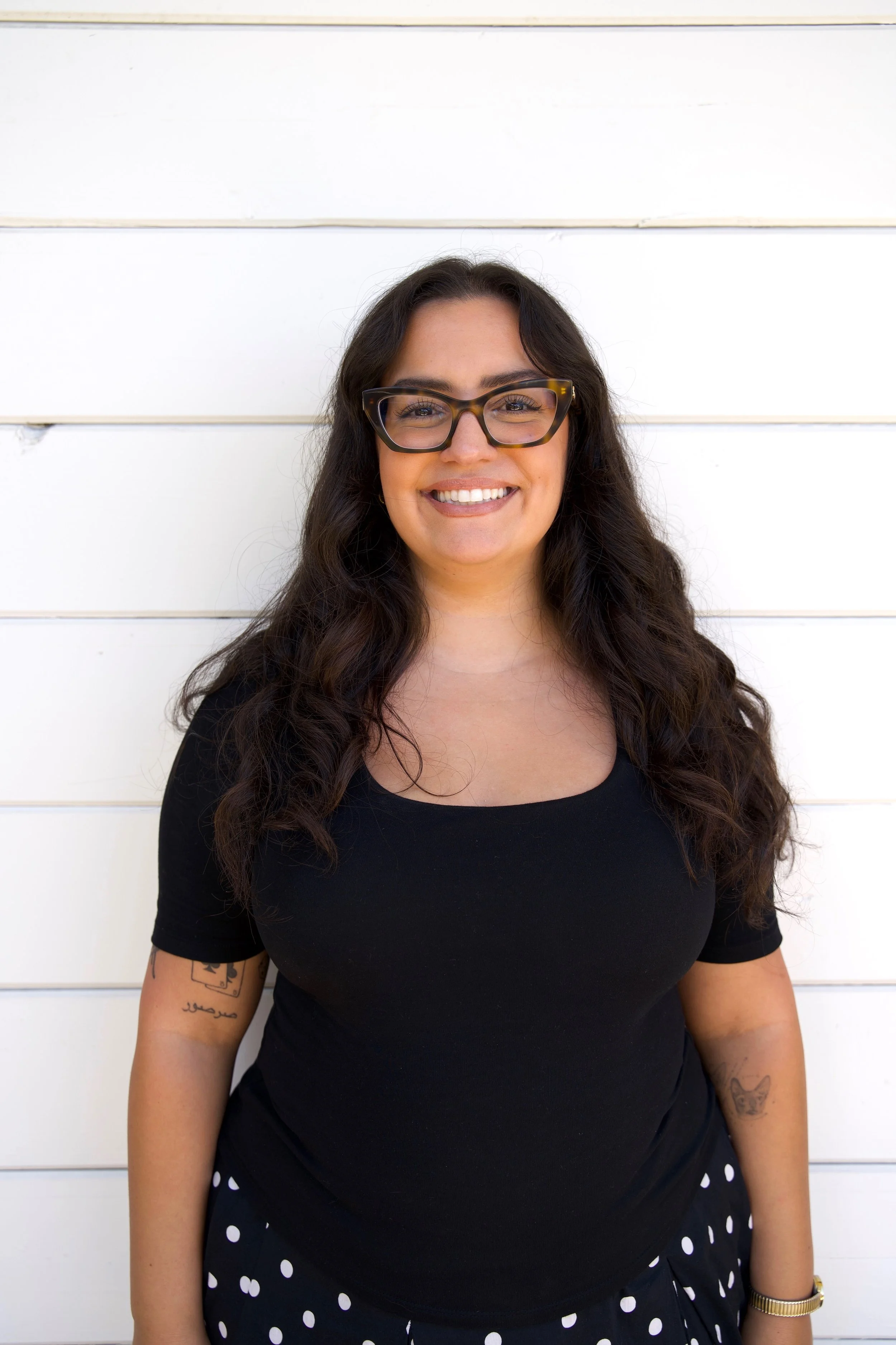 A woman with long dark hair, wearing glasses, a black top, and a polka dot skirt, smiling in front of a white wooden wall.