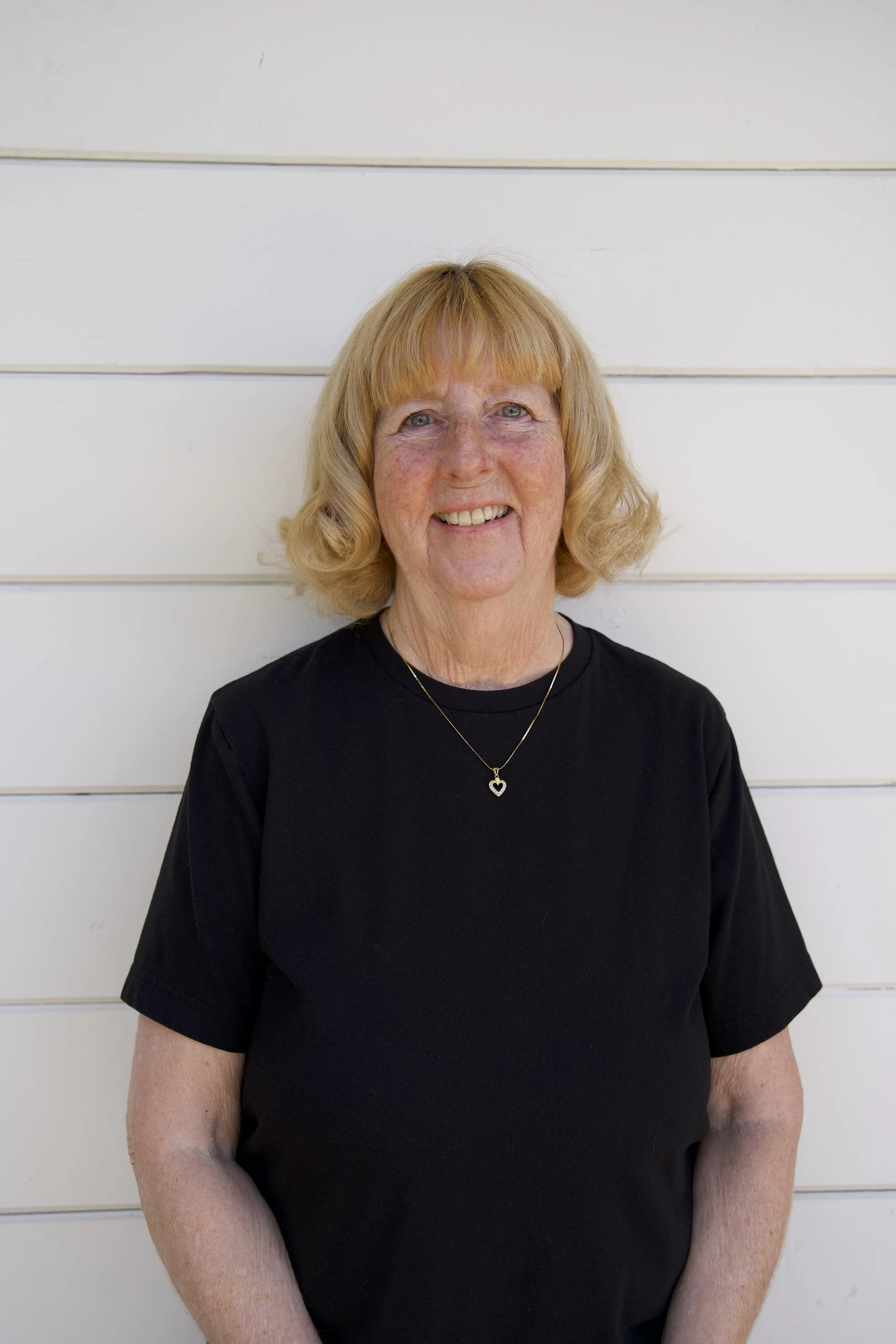 A smiling middle-aged woman with blonde hair wearing a black t-shirt and a necklace with a heart pendant standing in front of a white wooden wall.