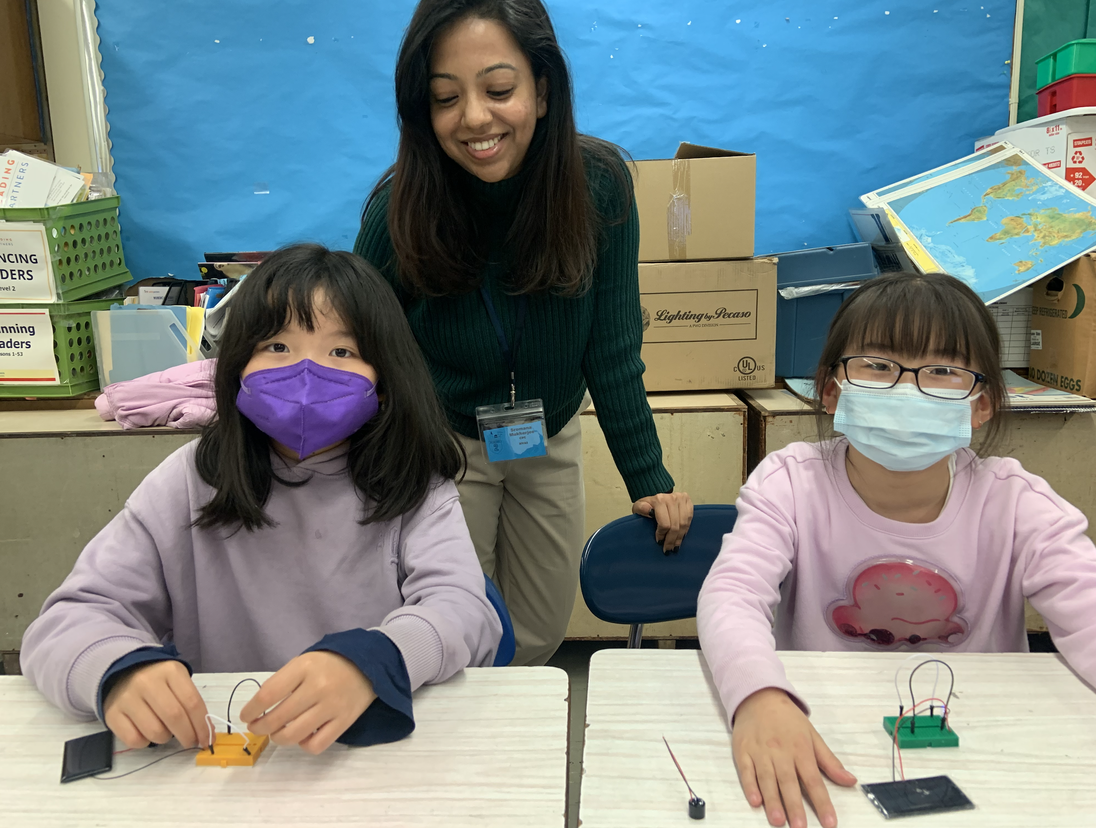 A woman and two young girls with masks engaged in a STEM activity at a classroom table. The woman is smiling and leaning over, while the girls are focused on their projects with wires and electronic components in front of them. Classroom supplies and a world map are visible in the background.