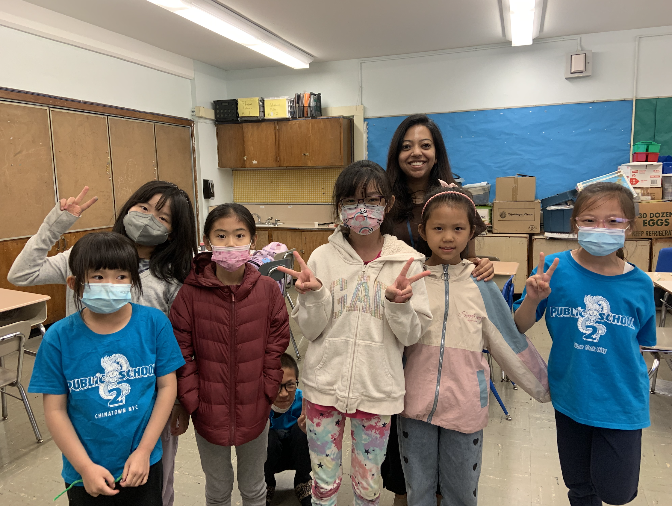 A group of children and a woman in a classroom, all wearing face masks and making peace signs.