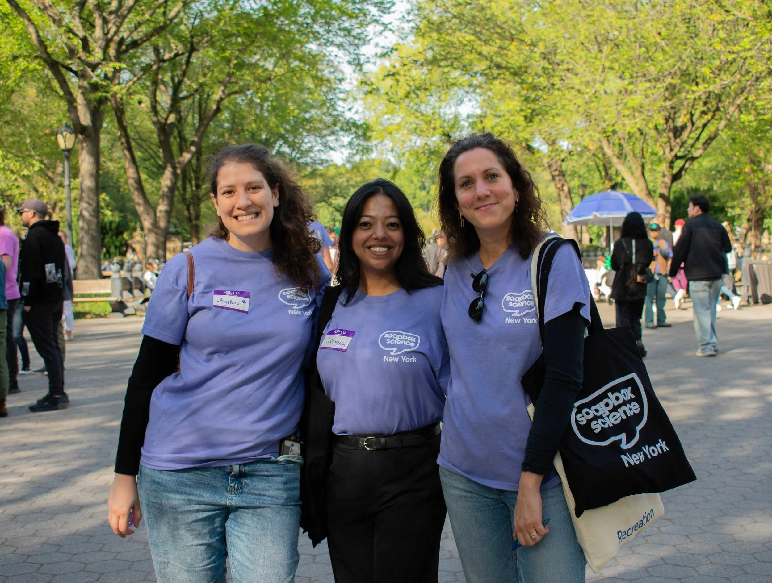 Three women standing together outdoors in a park, smiling at the camera, wearing purple 'Soapbox Science' t-shirts, with trees and people in the background.