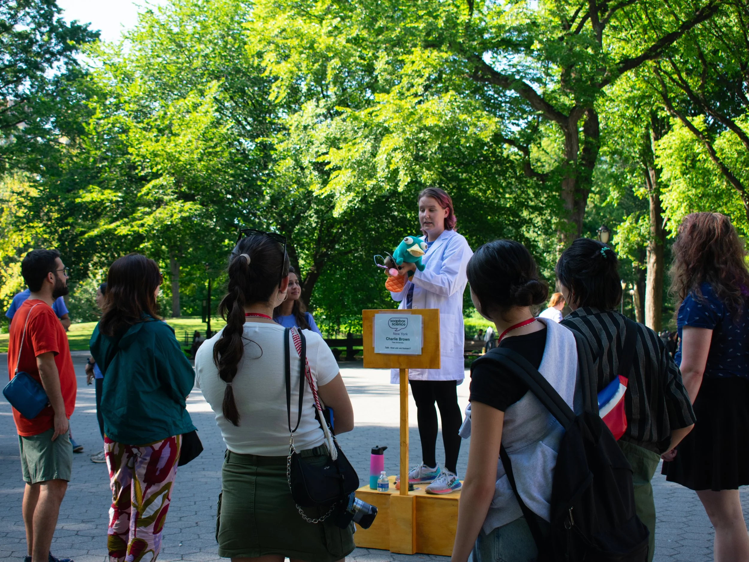 A woman in a white lab coat holding a puppet shows a group of people gathered around her outdoors in a park under green leafy trees.
