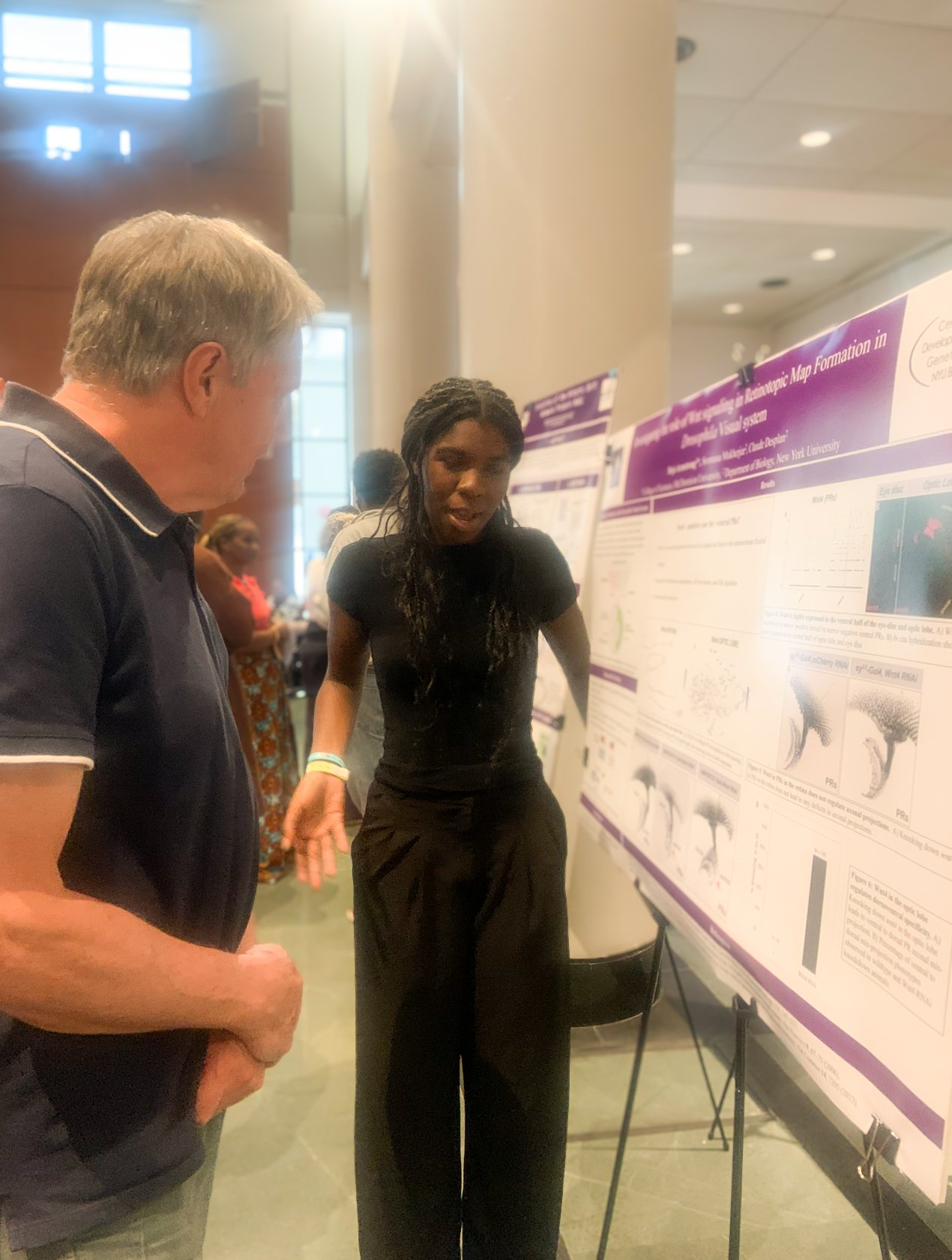 Two people engaged in discussion at a scientific research poster presentation. The woman is explaining her research to the man. The poster includes scientific diagrams and text.