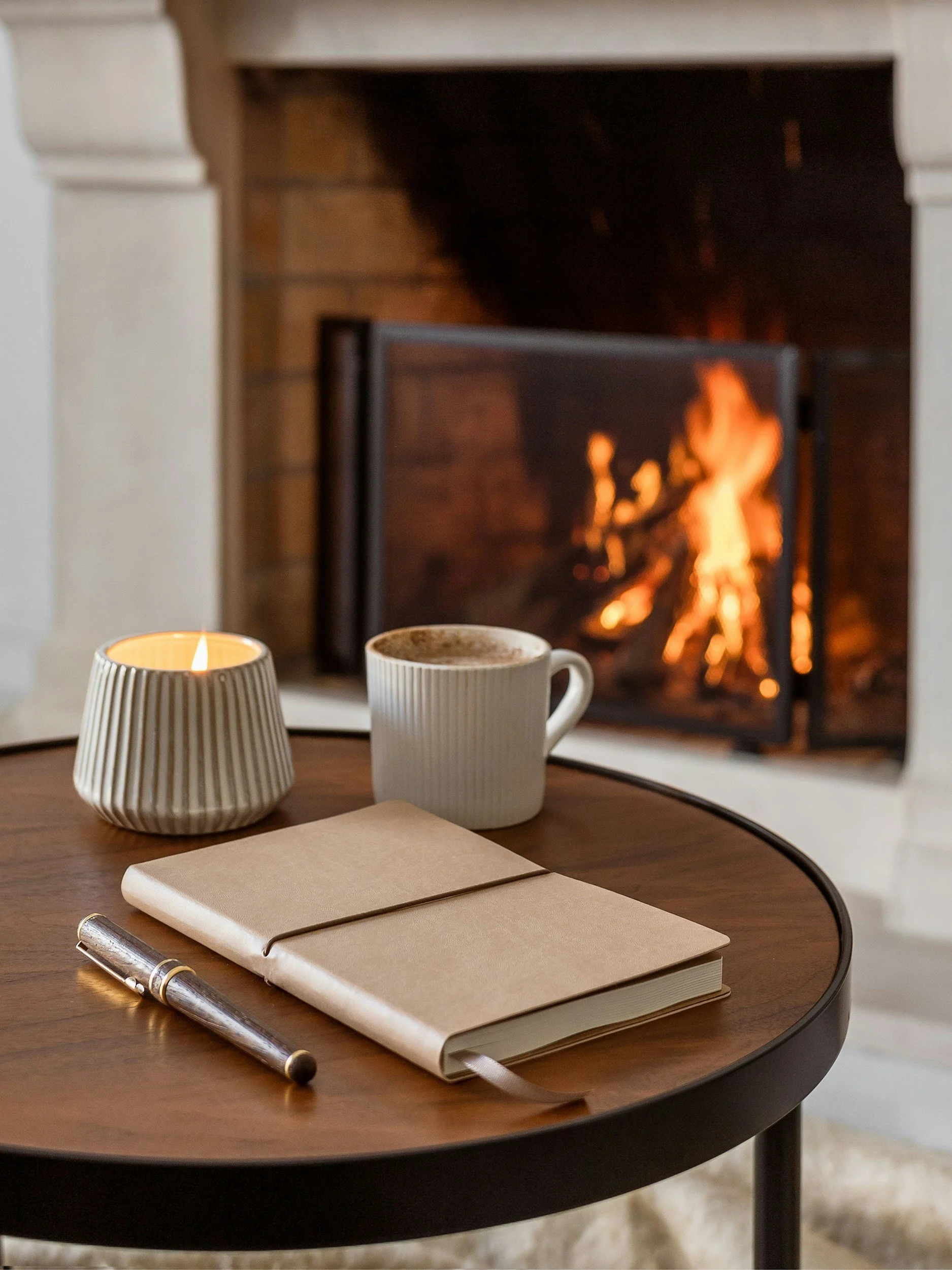 Cozy fireplace setting with a burning fire, a warm mug of cocoa, a journal, and a candle. A peaceful space illustrating morning devotionals, quiet rest, and mindset renewal for tired women