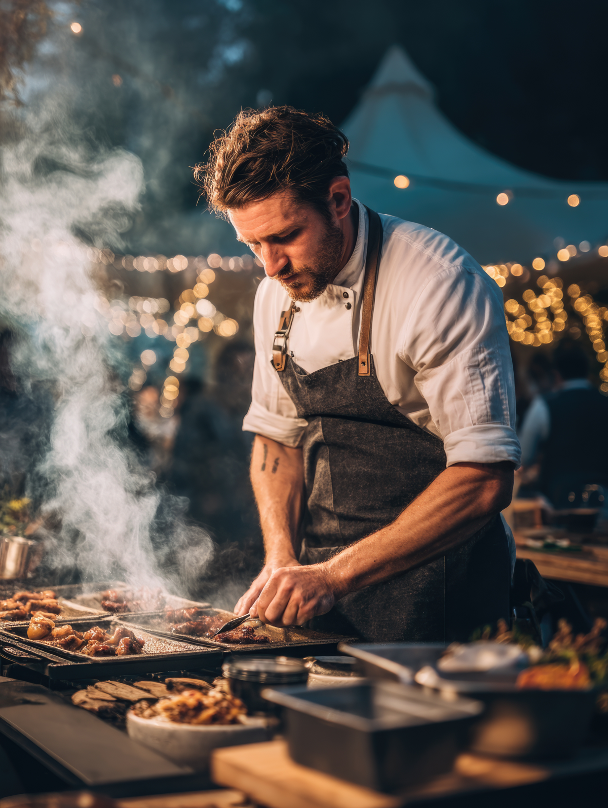 A male chef is cooking at an outdoor event during evening hours, with string lights and a white tent in the background, and steam rising from the grill.