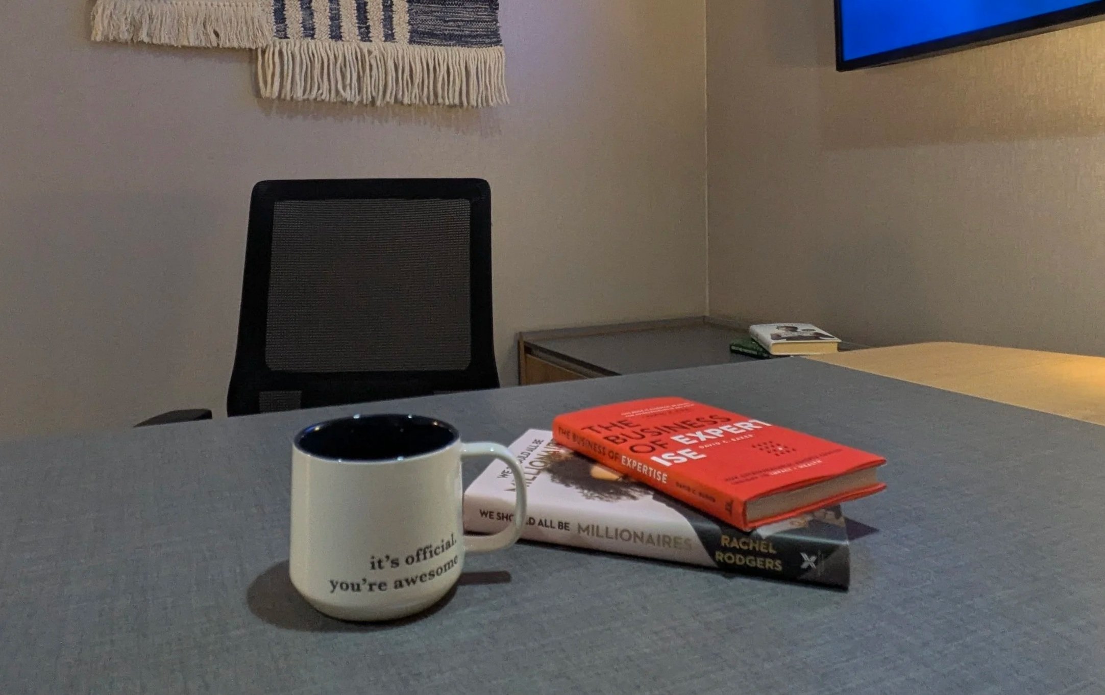 A coffee mug, two books, and a white notepad on a gray table in an office or conference room. A black mesh office chair is in the background, along with a wall hanging and a small cabinet. East Hartford Flex Space Private Office