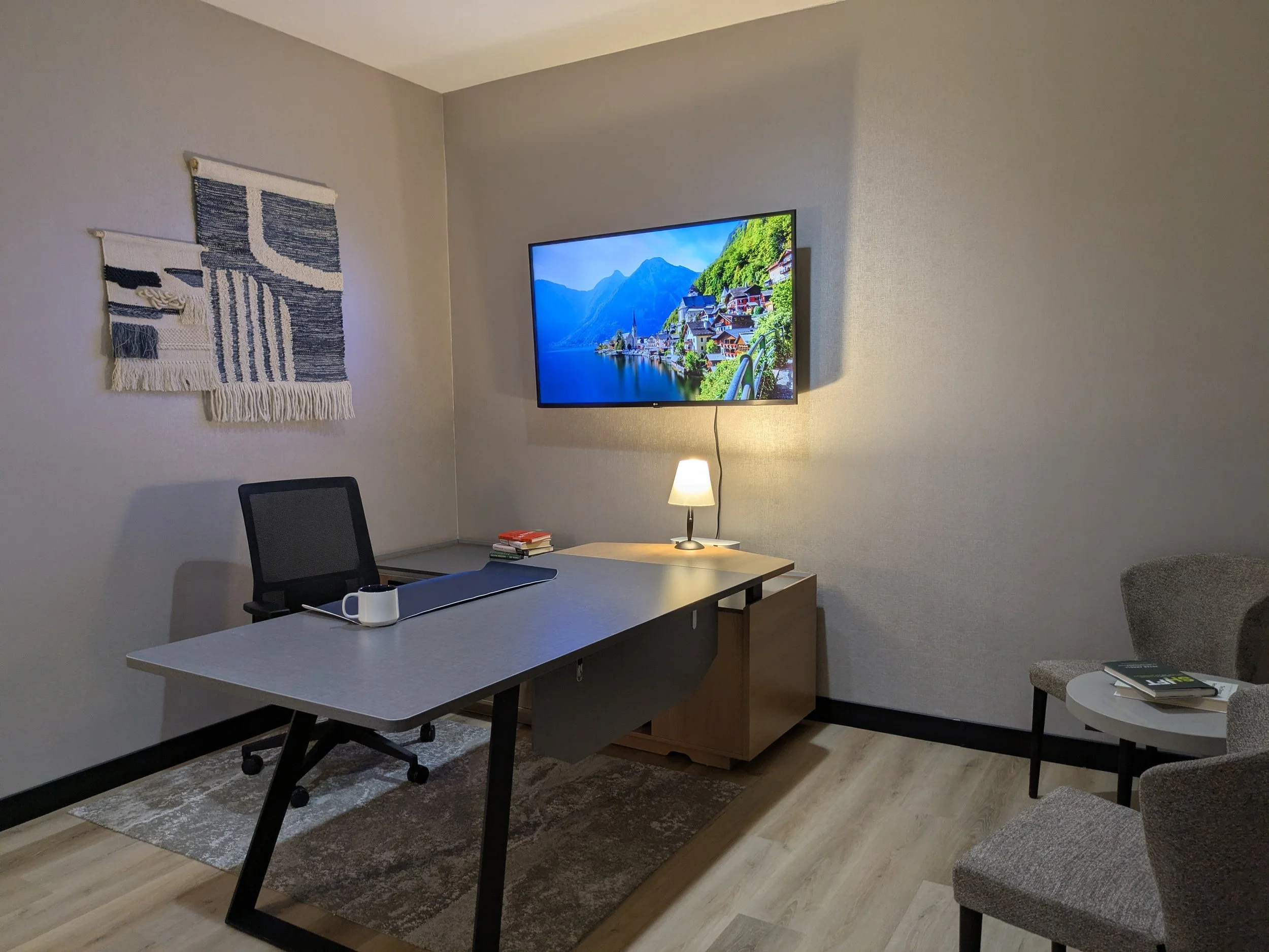 A modern home office with a desk, chair, wall art, a wall-mounted TV displaying a mountain and lake scene, a small lamp, and a side table with books. East Hartford Flex Space Coworking Private Office by the Hour Private Office by the Day