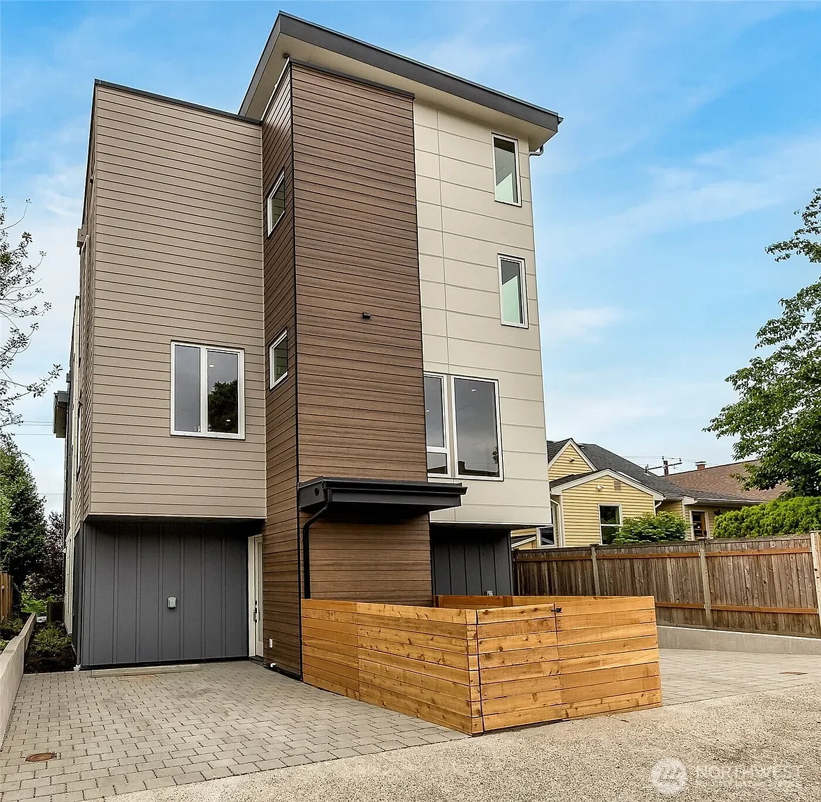 Modern multi-story house with beige and brown exterior siding, large windows, and a small wooden planter box in the front yard, set against a blue sky.