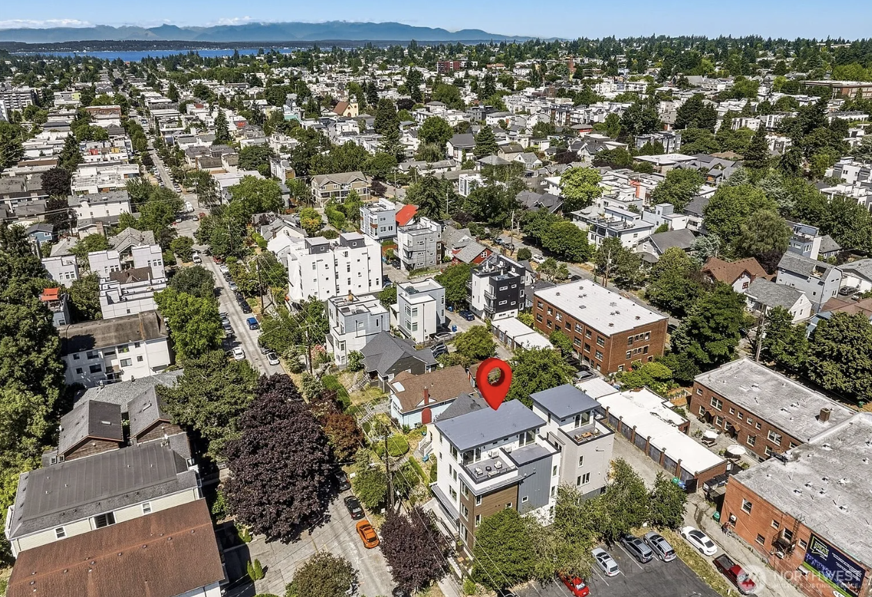 An aerial view of a residential neighborhood with various houses, trees, and parking lots, extending to a distant body of water and distant mountains under a blue sky.