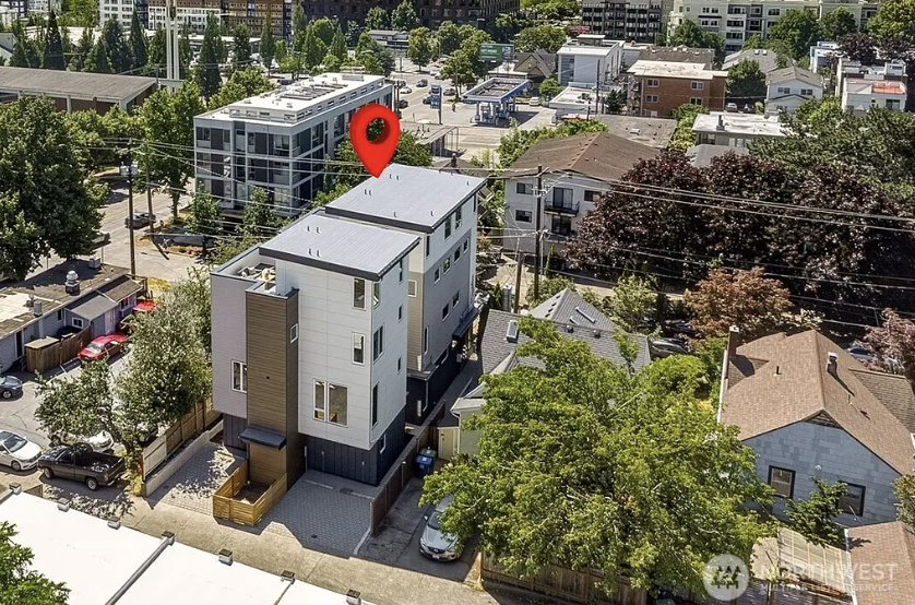 Aerial view of a modern multi-story building with a flat roof, surrounded by trees and neighboring houses in an urban area. A red location marker is positioned over the building.