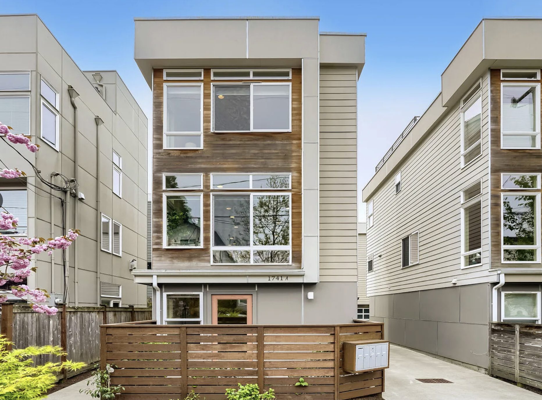 Front of a modern multi-story residential building with large windows and wood paneling accents, neighboring similar buildings on either side, a small outdoor area with a wooden fence, plants, and mailboxes in the foreground, and clear blue sky above