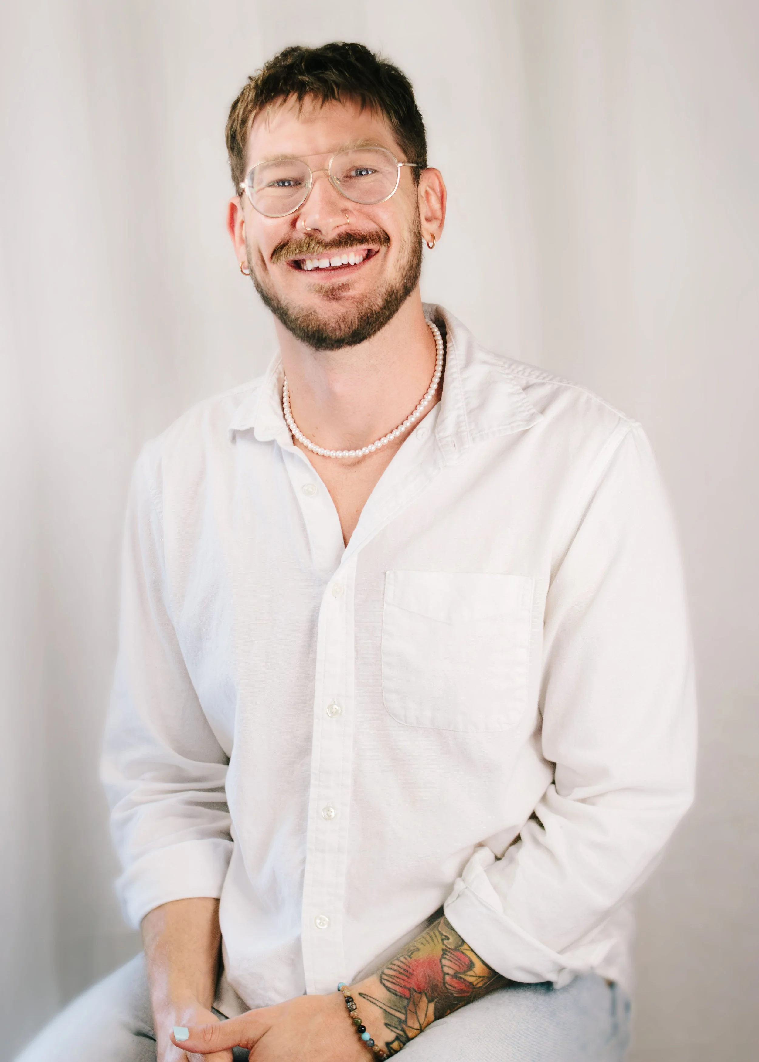 A young man with short dark hair, glasses, and a beard, smiling, wearing a white button-up shirt, necklace, earrings, and a colorful tattoo on his arm. He is sitting against a plain white background.