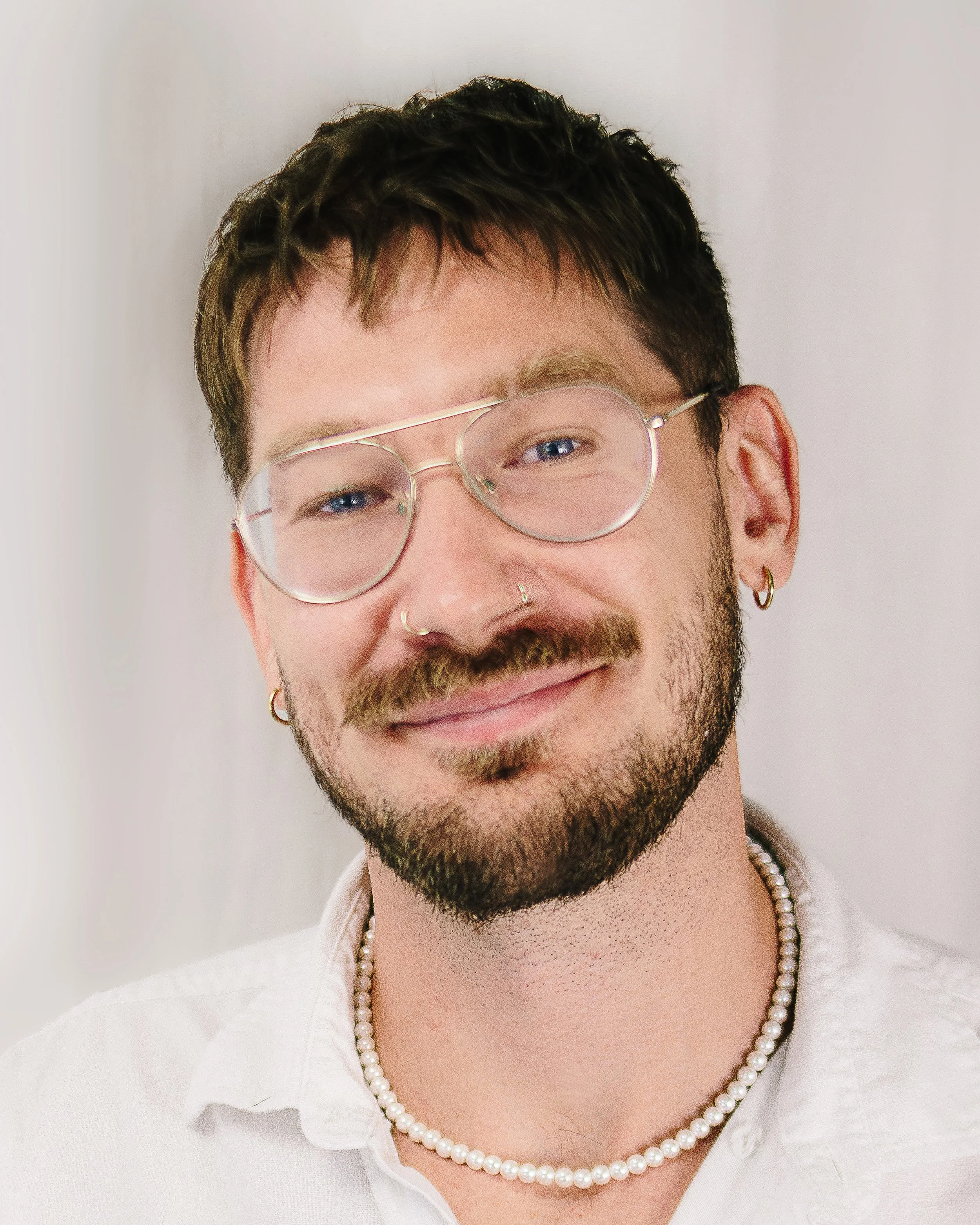 Close-up of a man with short dark hair, glasses, earrings, a nose ring, a pearl necklace, and a white shirt, smiling.