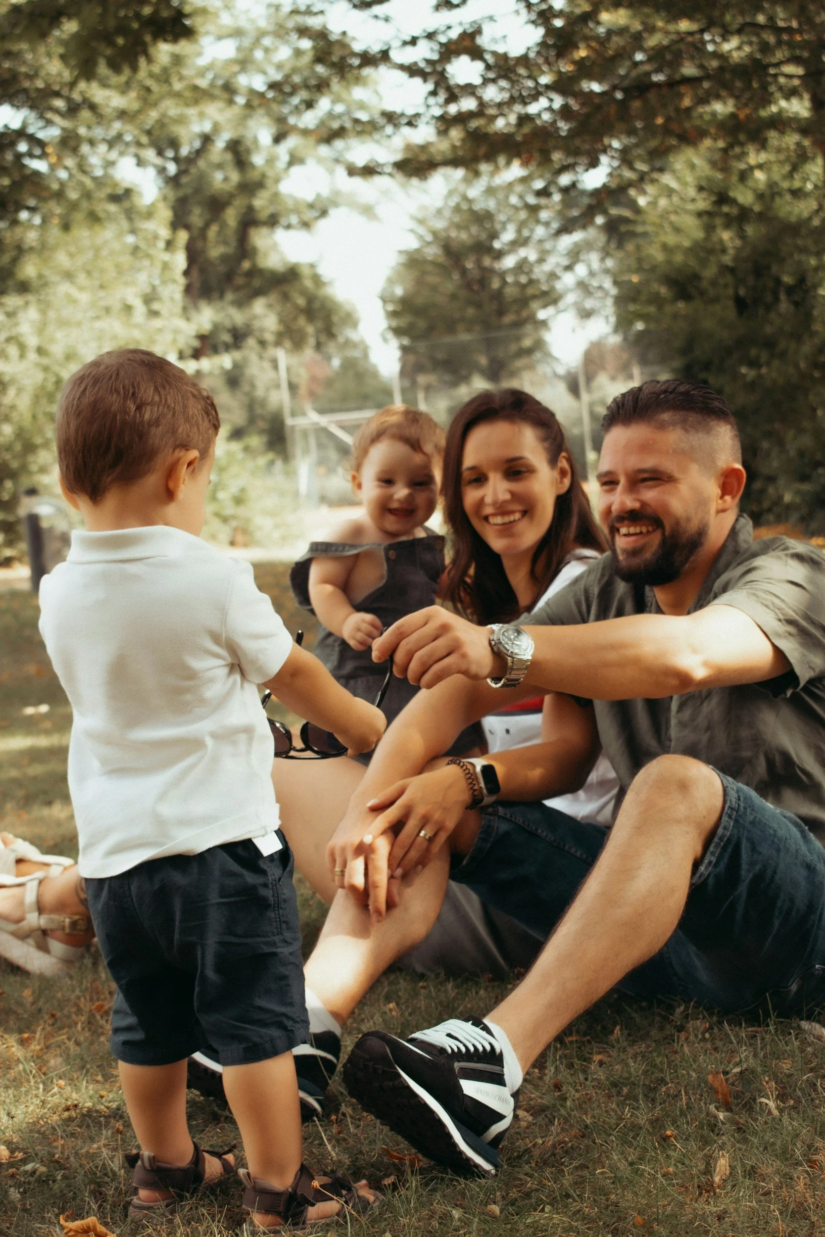 A family of four outdoors on a sunny day, with three children and two adults, sitting on grass beneath trees. One child faces away from the camera, while the other child and the two adults face and smile at each other.