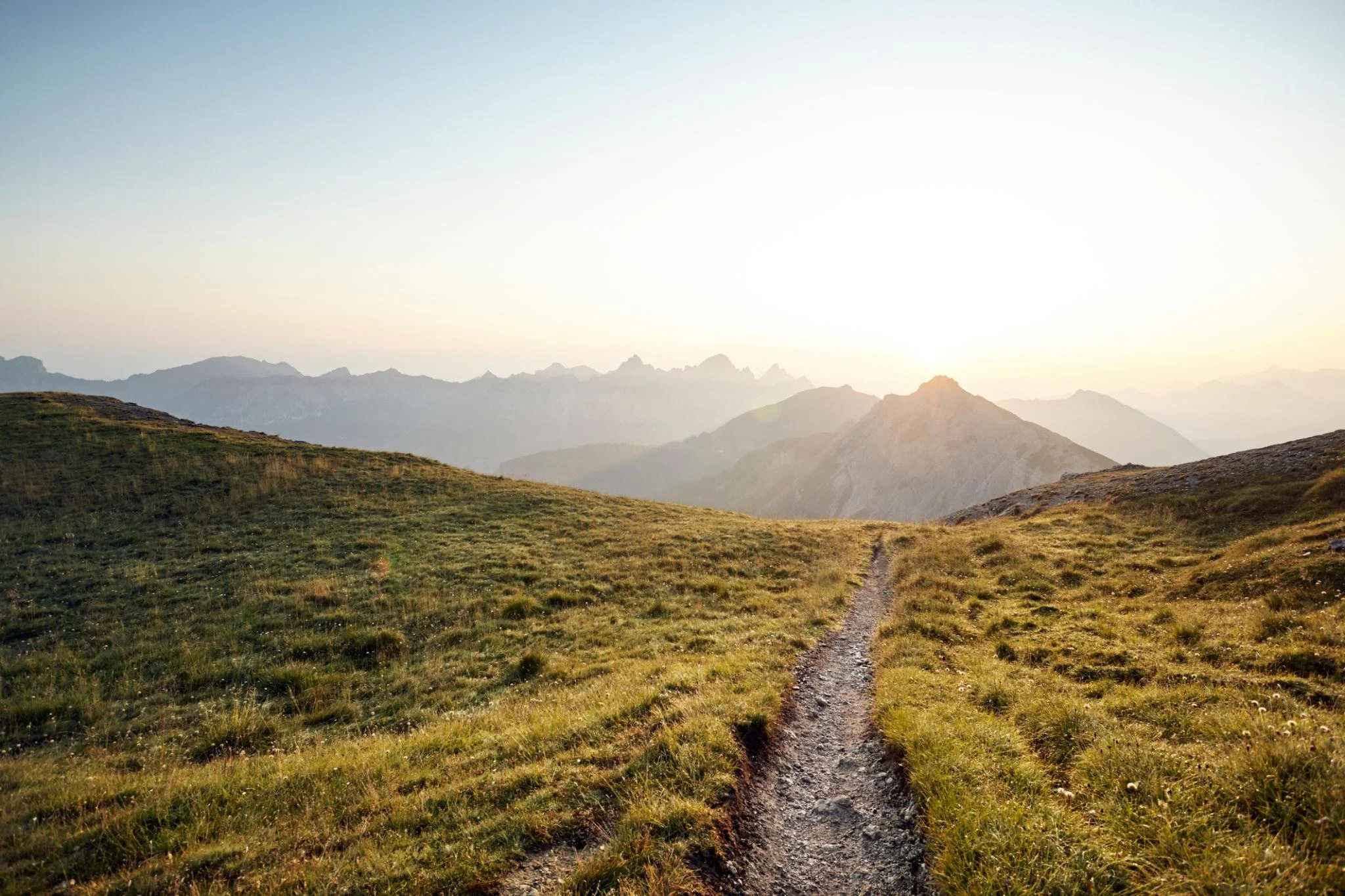 A dirt path winds through green grassy fields with mountain peaks in the distance under a clear sky at sunrise or sunset.