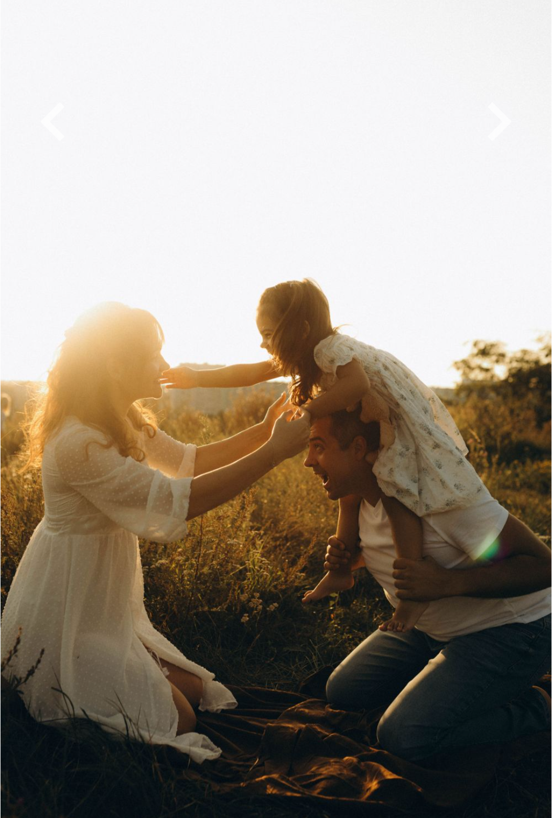 A family of four enjoying a playful moment outdoors during sunset. The mother and father are on their knees, with their young daughter sitting on the father's shoulders. The daughter is reaching out to touch her mother's face, and everyone appears happy and engaged in a family activity.