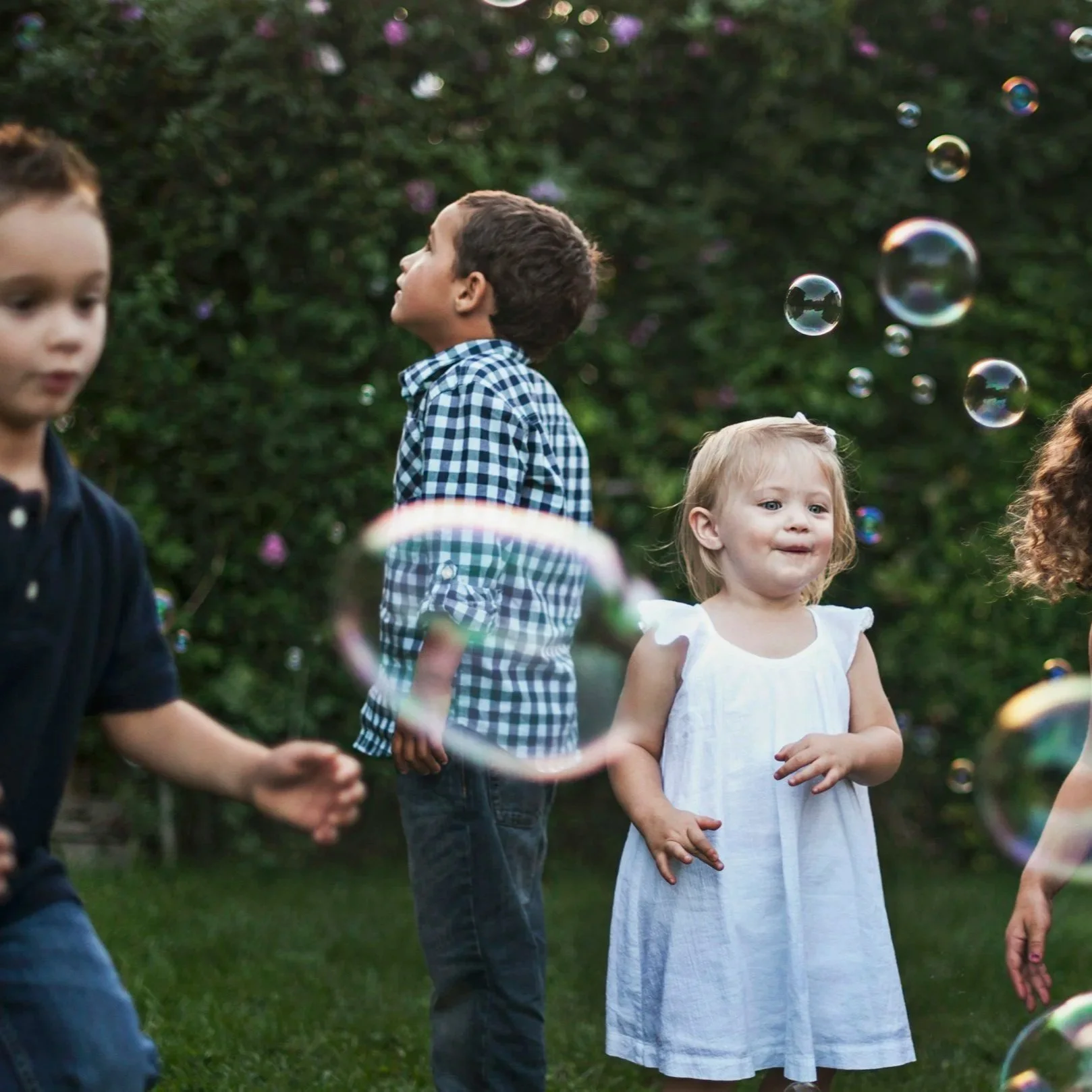 Children playing with bubbles outdoors, with a green bush background.