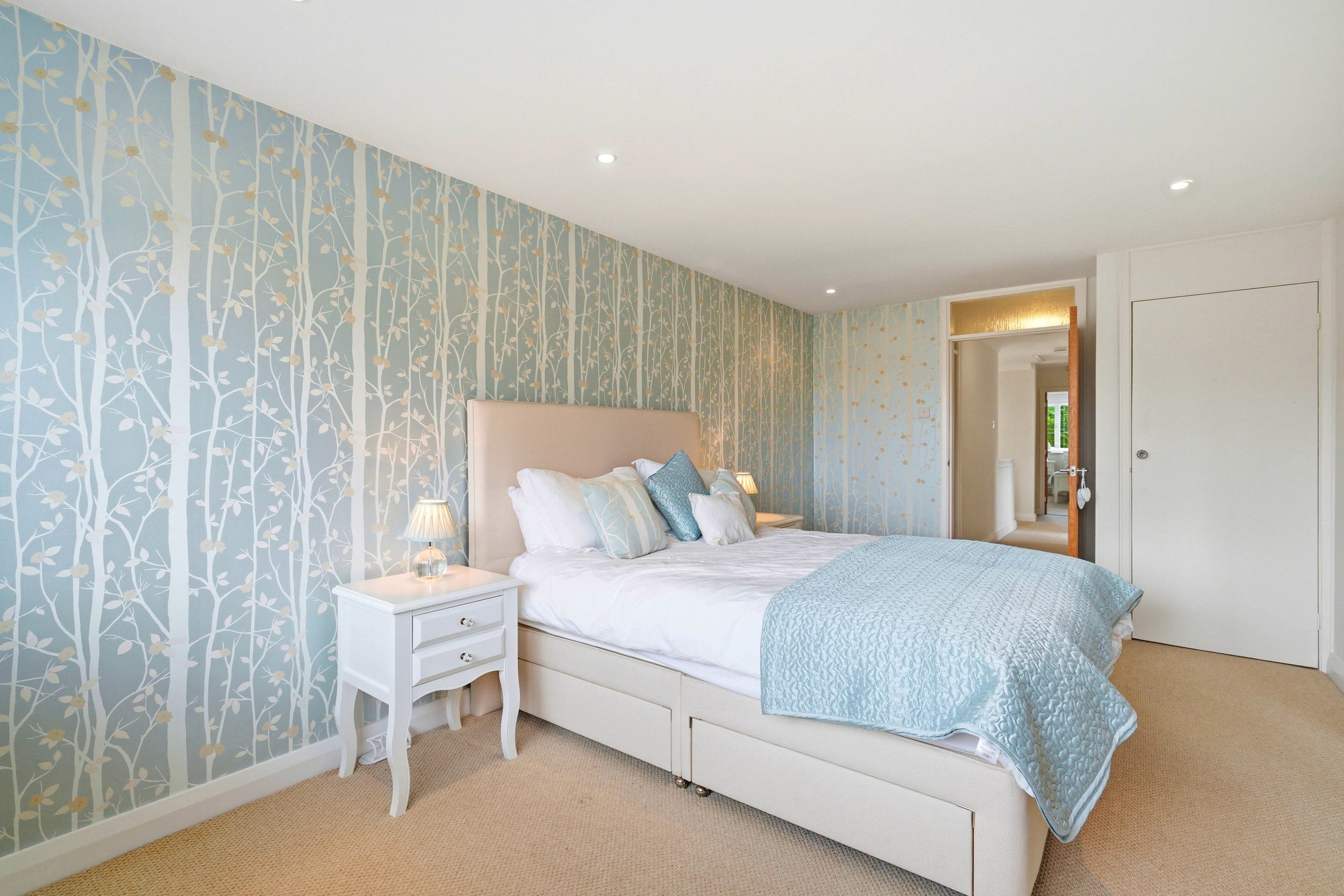 A bedroom with a bed, nightstand, and wallpapered accent wall featuring a tree and leaf pattern in light blue and gold tones.