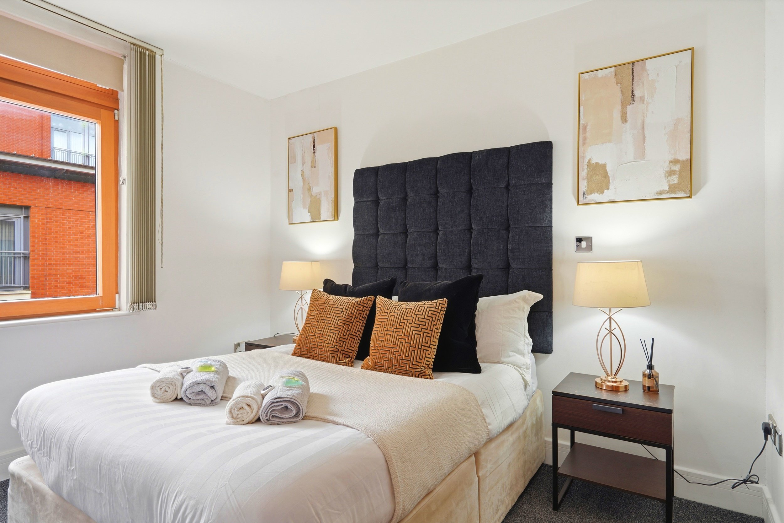 A bedroom with a large bed covered with a white striped duvet, six rolled towels on it, and four decorative pillows with orange and black patterns. The bed has a tall dark gray tufted headboard. Next to the bed is a dark wooden nightstand with a rose