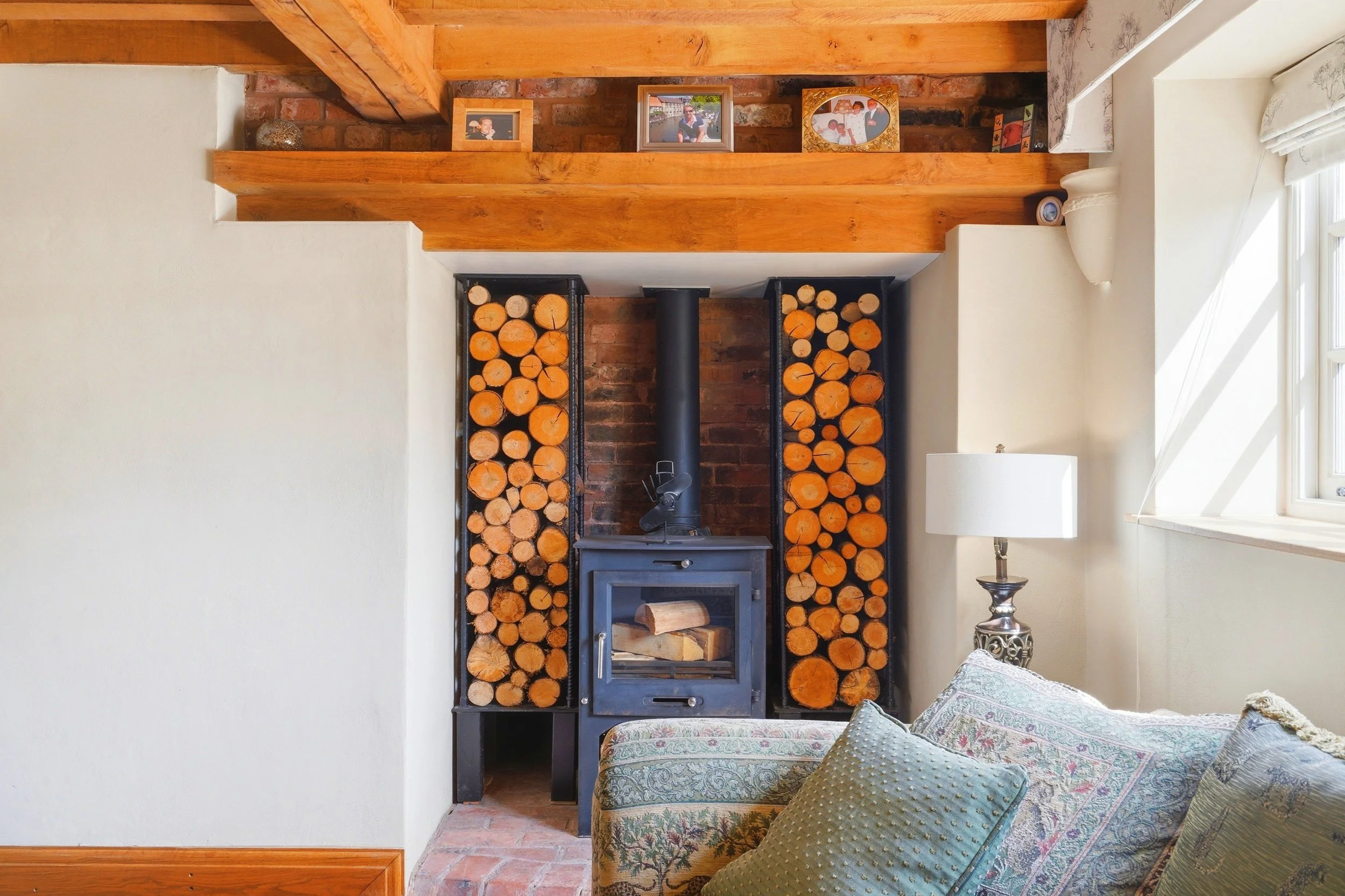 Indoor living room with a wood-burning stove, stacked firewood on each side, brick and wooden accents, and a floral-patterned sofa with pillows.