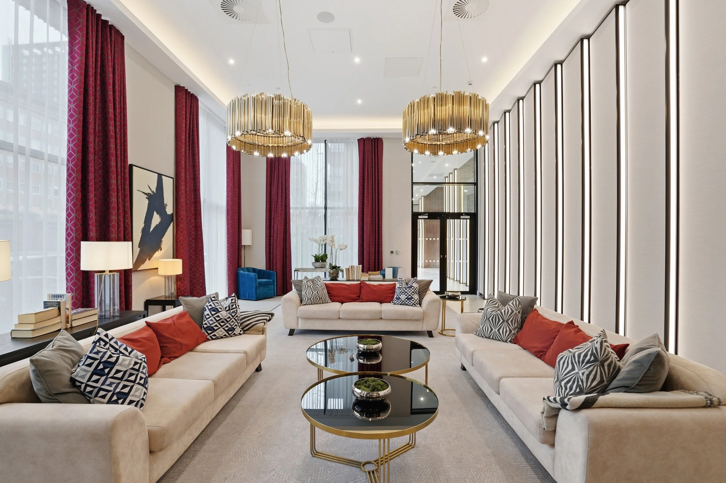 A modern, upscale living room with beige sofas, decorative pillows, glass-top coffee tables, red curtains, contemporary artwork, and large chandeliers.