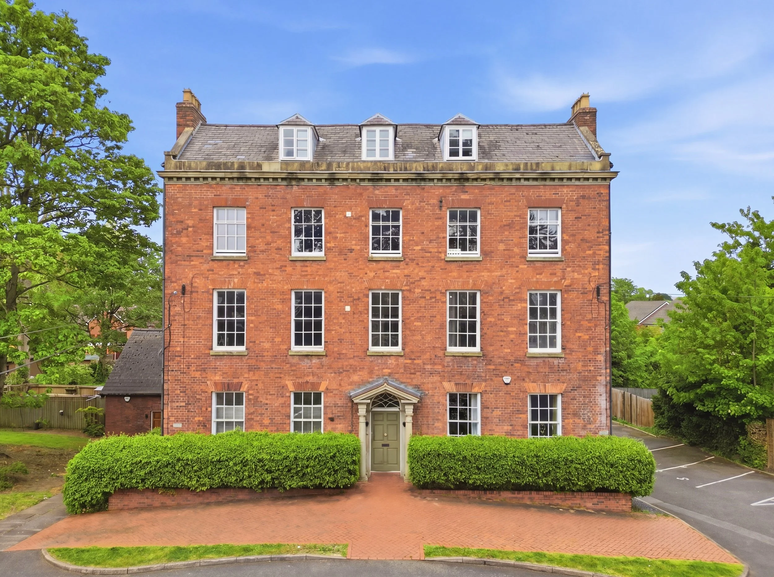 A red brick multi-story building with white window frames and a green front door. It is surrounded by green bushes and trees, with a blue sky in the background.