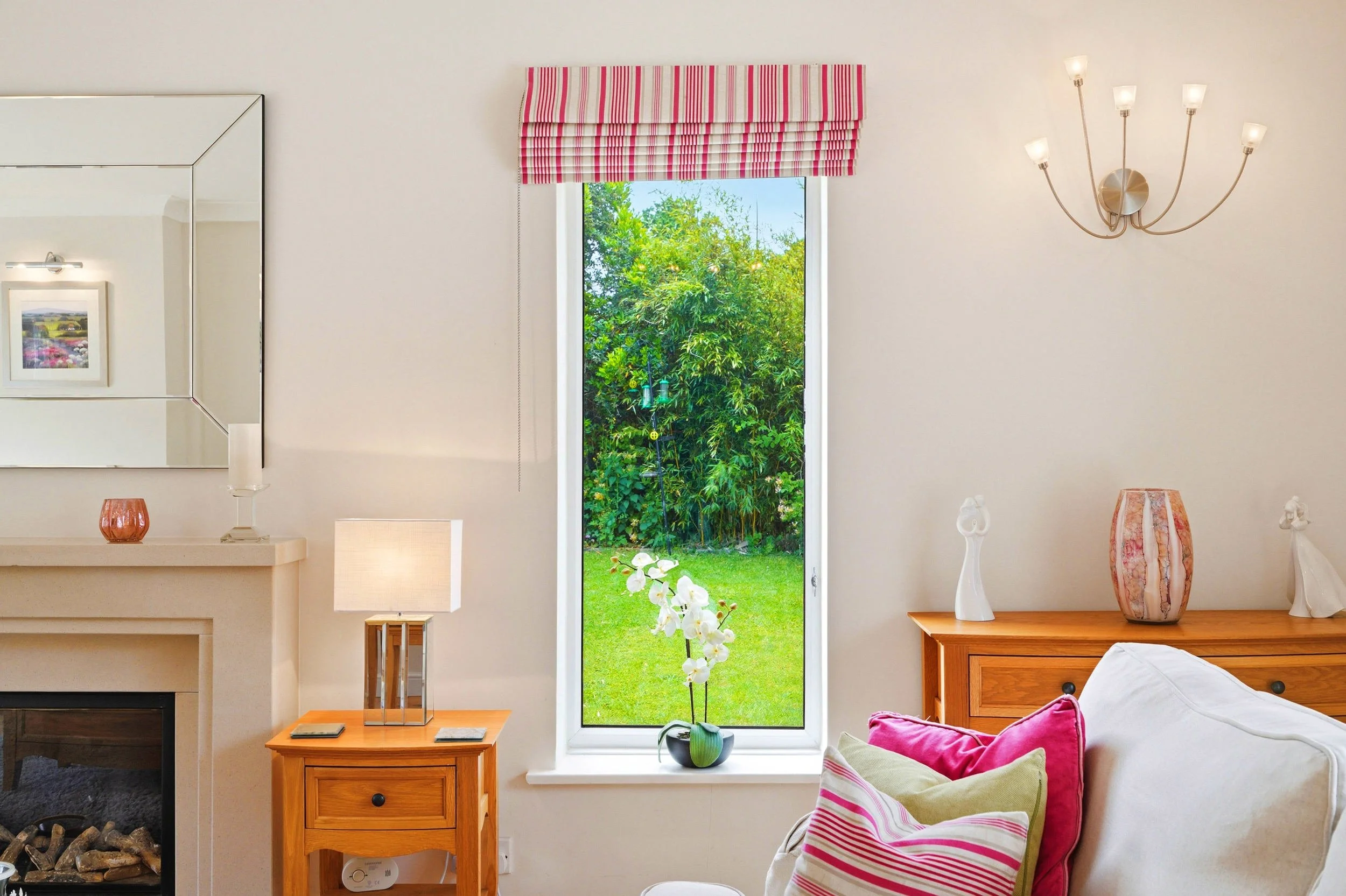 A cozy living room with a large window showing a lush green garden outside. Inside, there is a white wall with a decorative sconce light, a mirror, and a small wooden side table with a lamp and books. On the right, a wooden cabinet holds vases and de