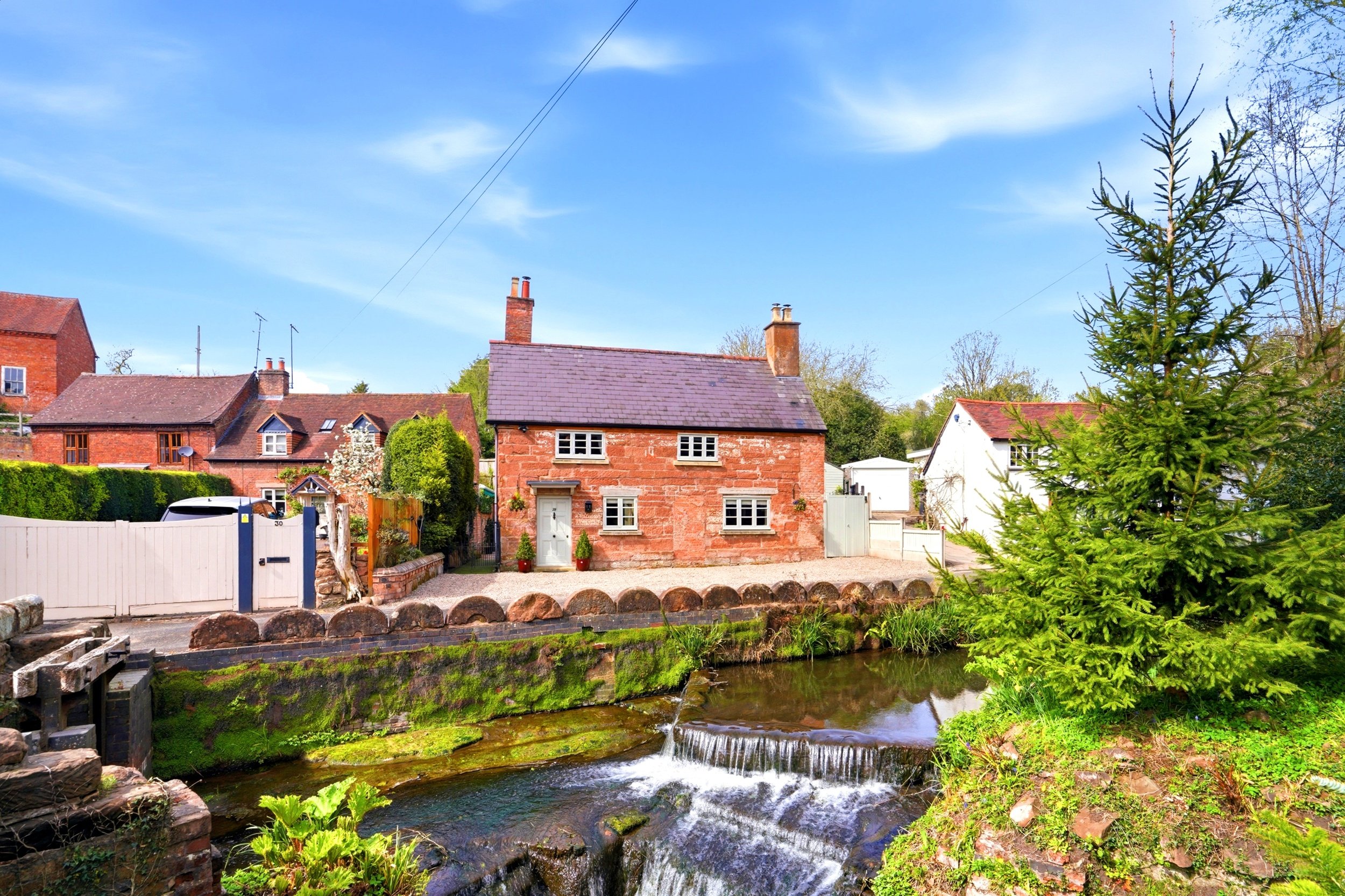 View of a small red brick house beside a creek, with greenery, trees, and a blue sky with scattered clouds in a rural setting.