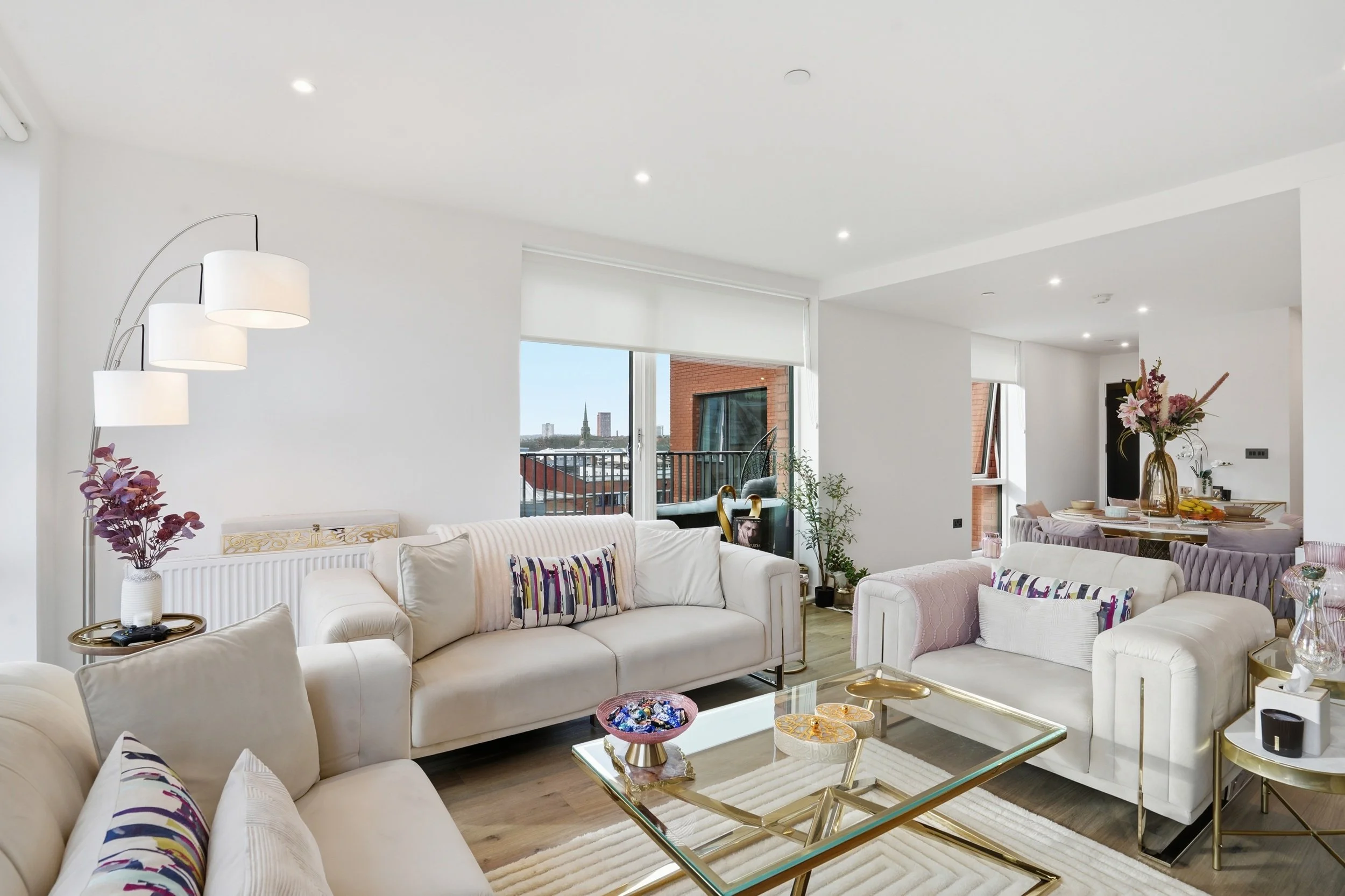 Modern living room with white sofas, glass coffee table, tall white curtains, and large windows showing city buildings. Decor includes a tall arc floor lamp with multiple lampshades, potted plants, and a dining area with a flower vase and tableware.