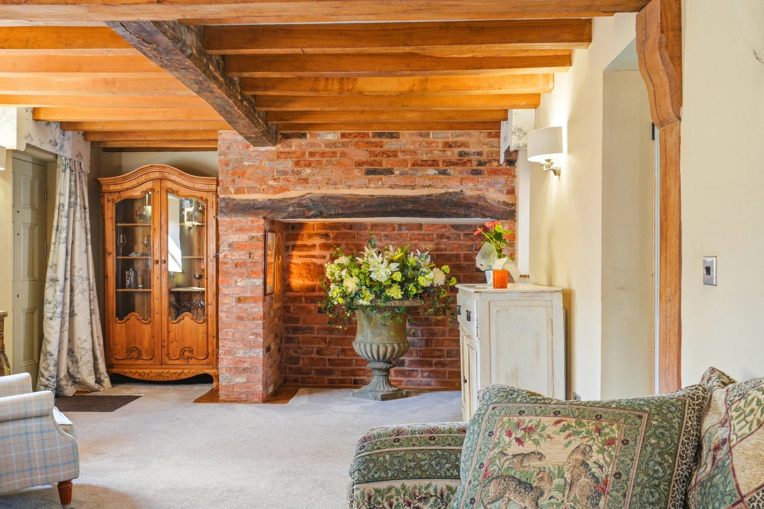 Living room with brick fireplace, wooden beams, and floral furniture, with a floral arrangement on a large urn.