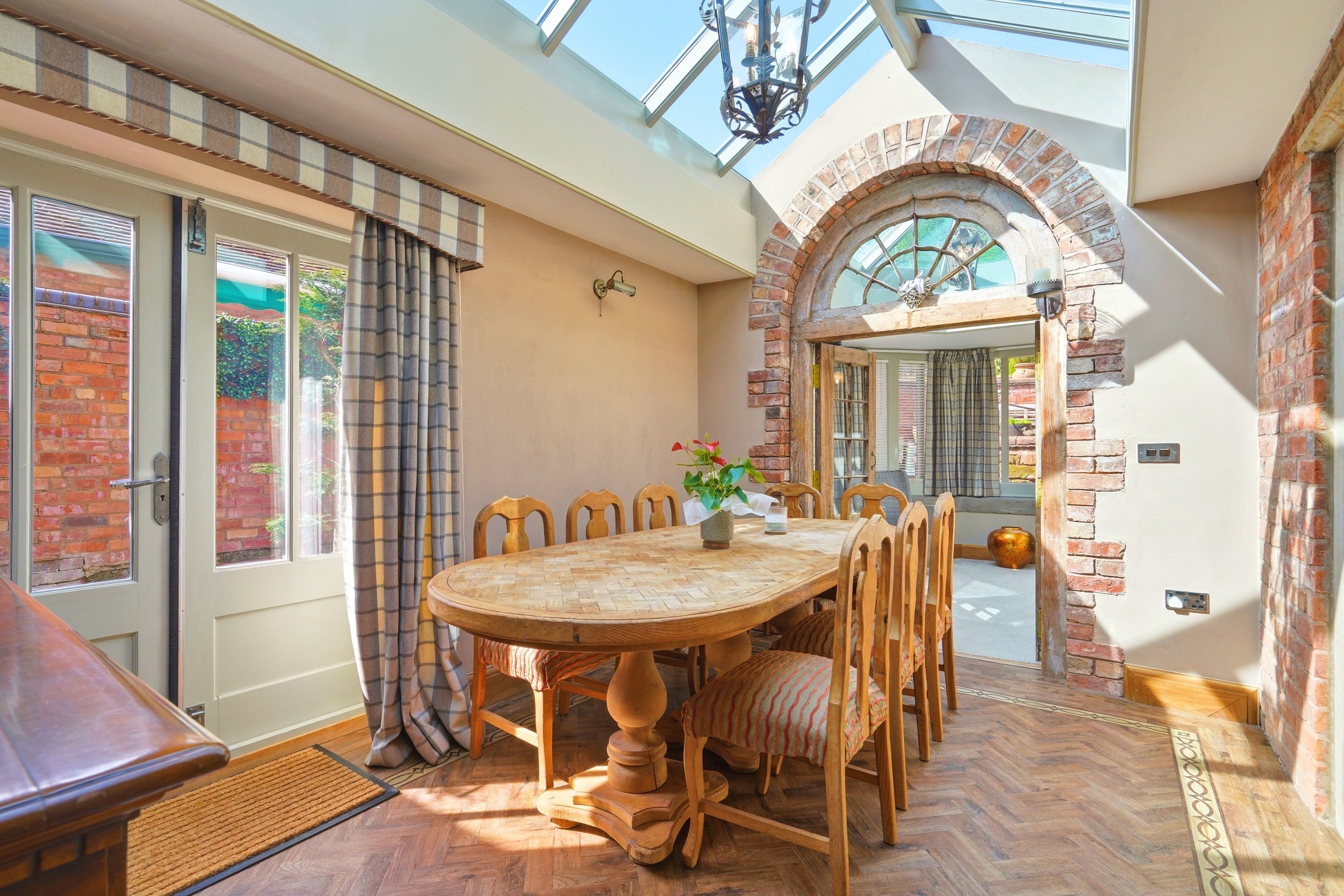 A bright dining room features a wooden table with chairs, brick wall accents, large windows with plaid curtains, and a glass ceiling letting in natural light.