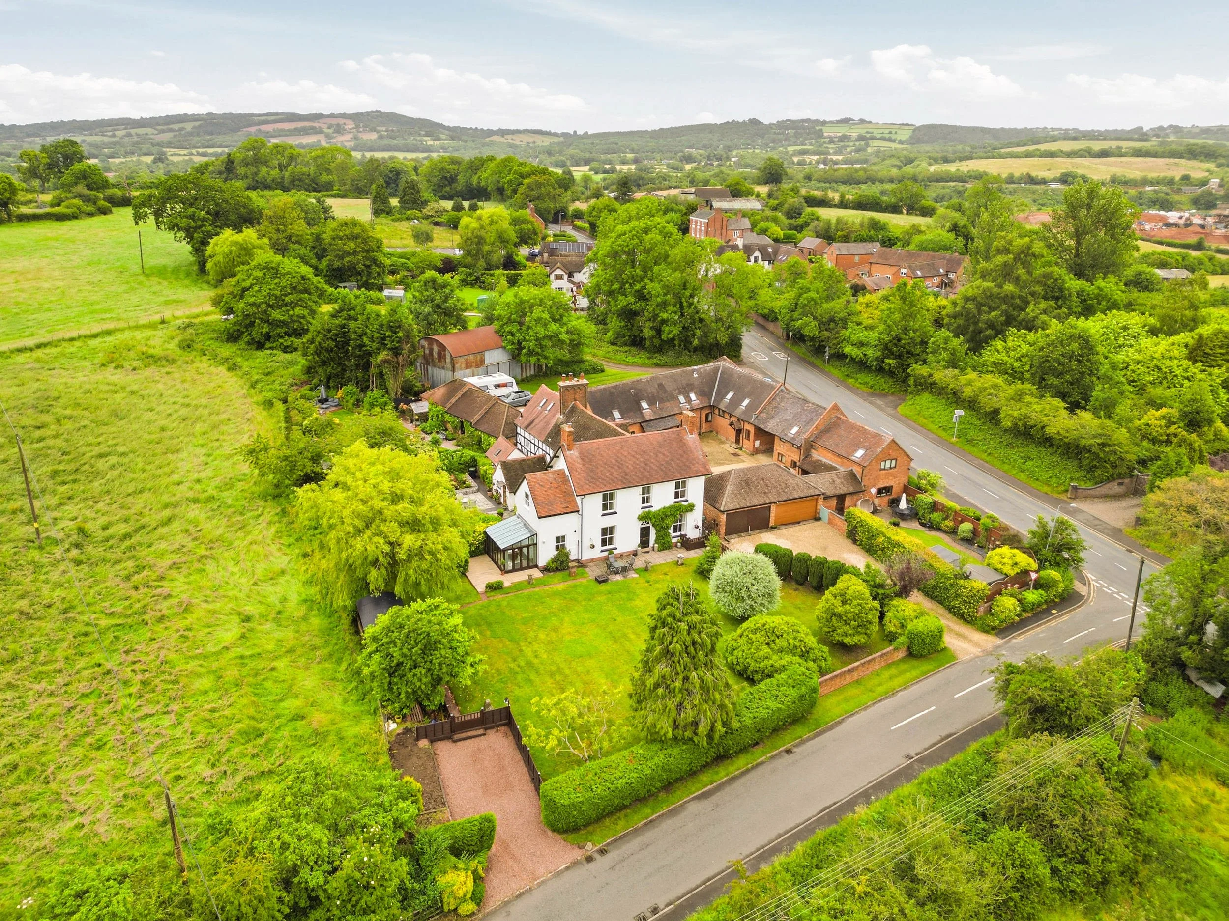 A panoramic aerial view of a suburban neighborhood surrounded by lush green trees and fields, with houses, roads, and rolling hills in the background on a partly cloudy day.