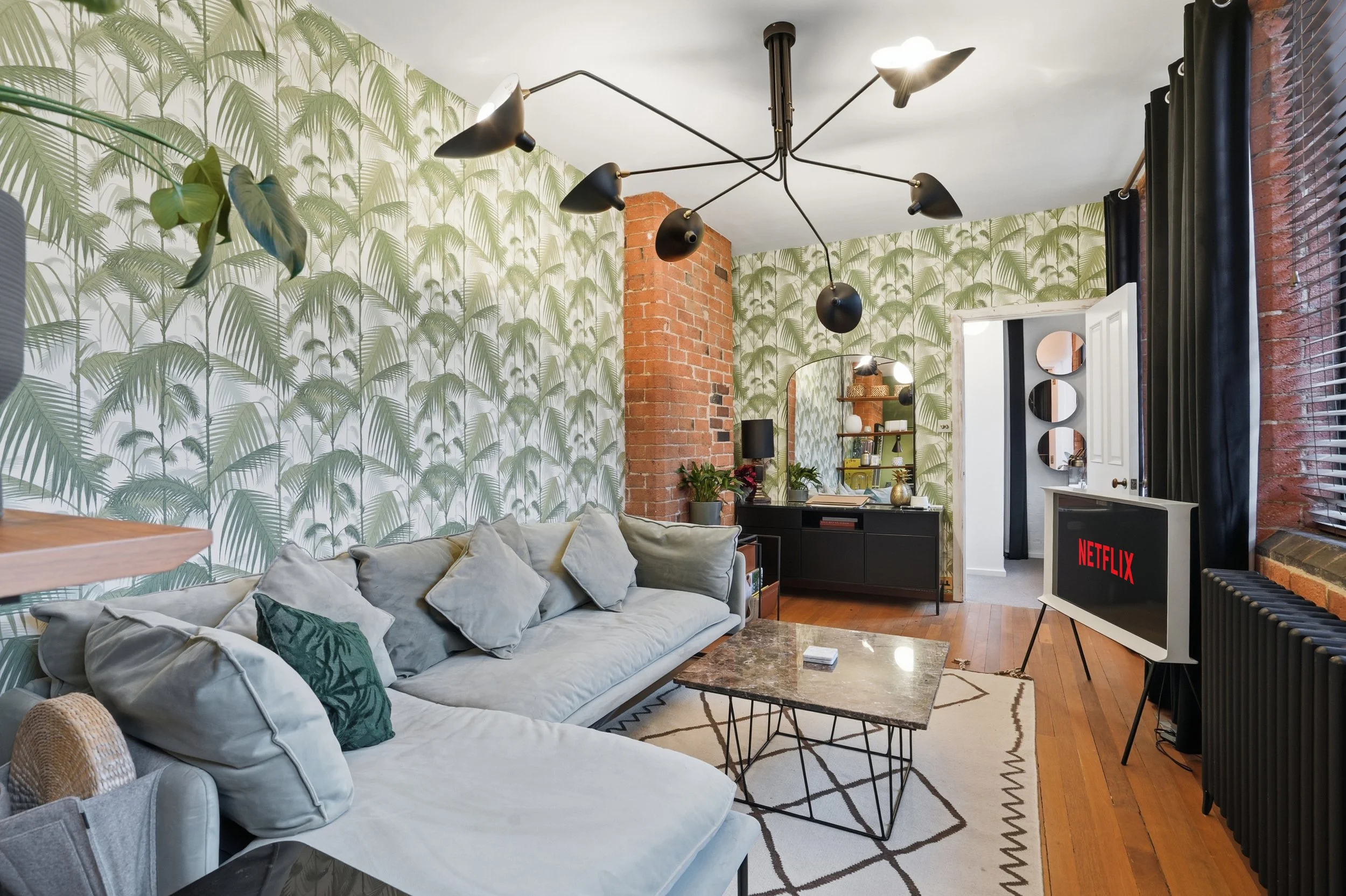 Living room with green palm leaf wallpaper, brick pillars, black modern chandelier, beige sofa with pillows, black cabinet, bookshelf with plants and decor, TV displaying Netflix, area rug, black curtains, hardwood floor.