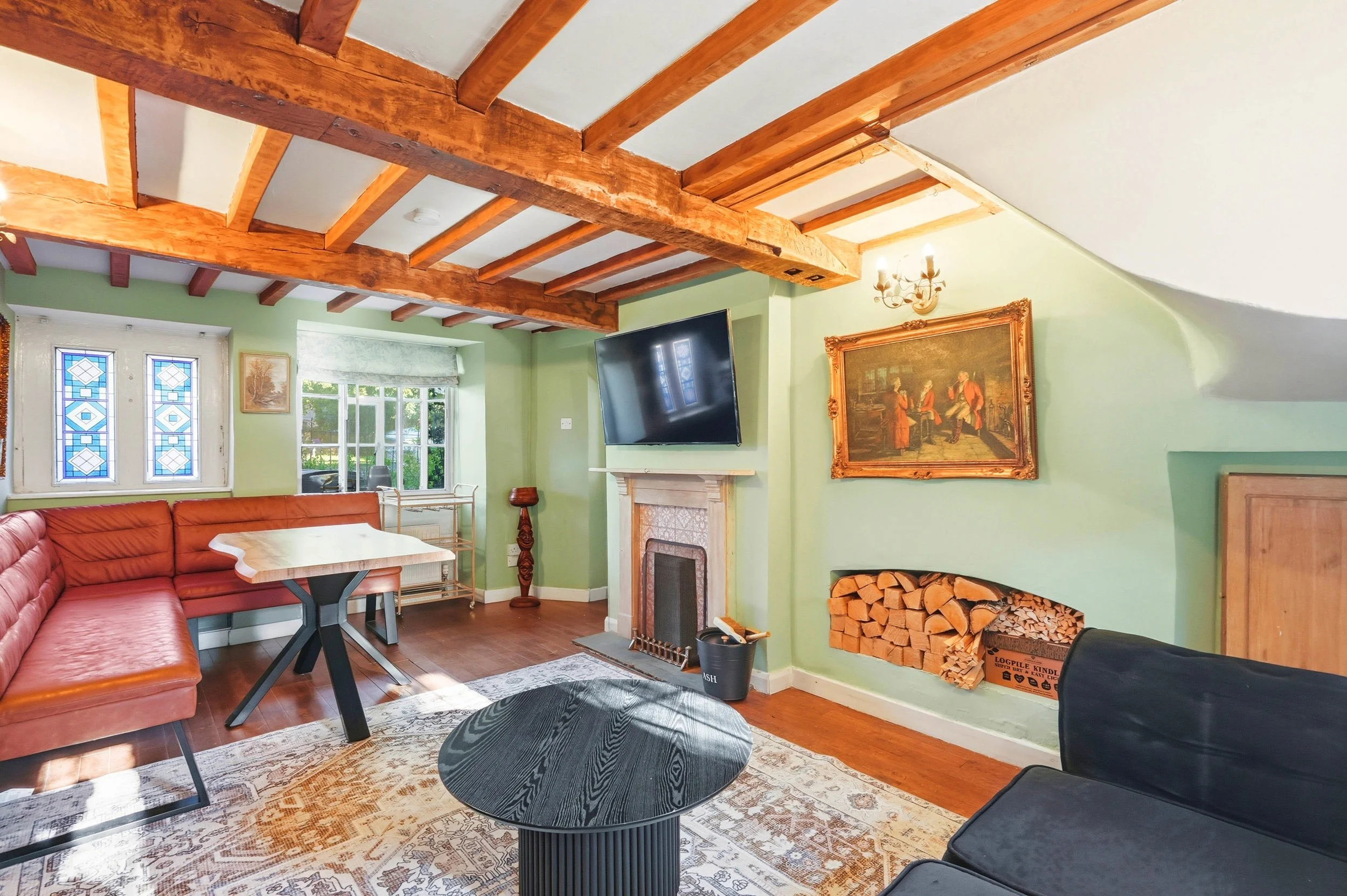 Living room with green walls, wooden ceiling beams, a brick fireplace with a TV mounted above, and a woodpile stored in a built-in corner. There is a red sectional sofa, a black coffee table, and a patterned area rug. The room has natural light comin