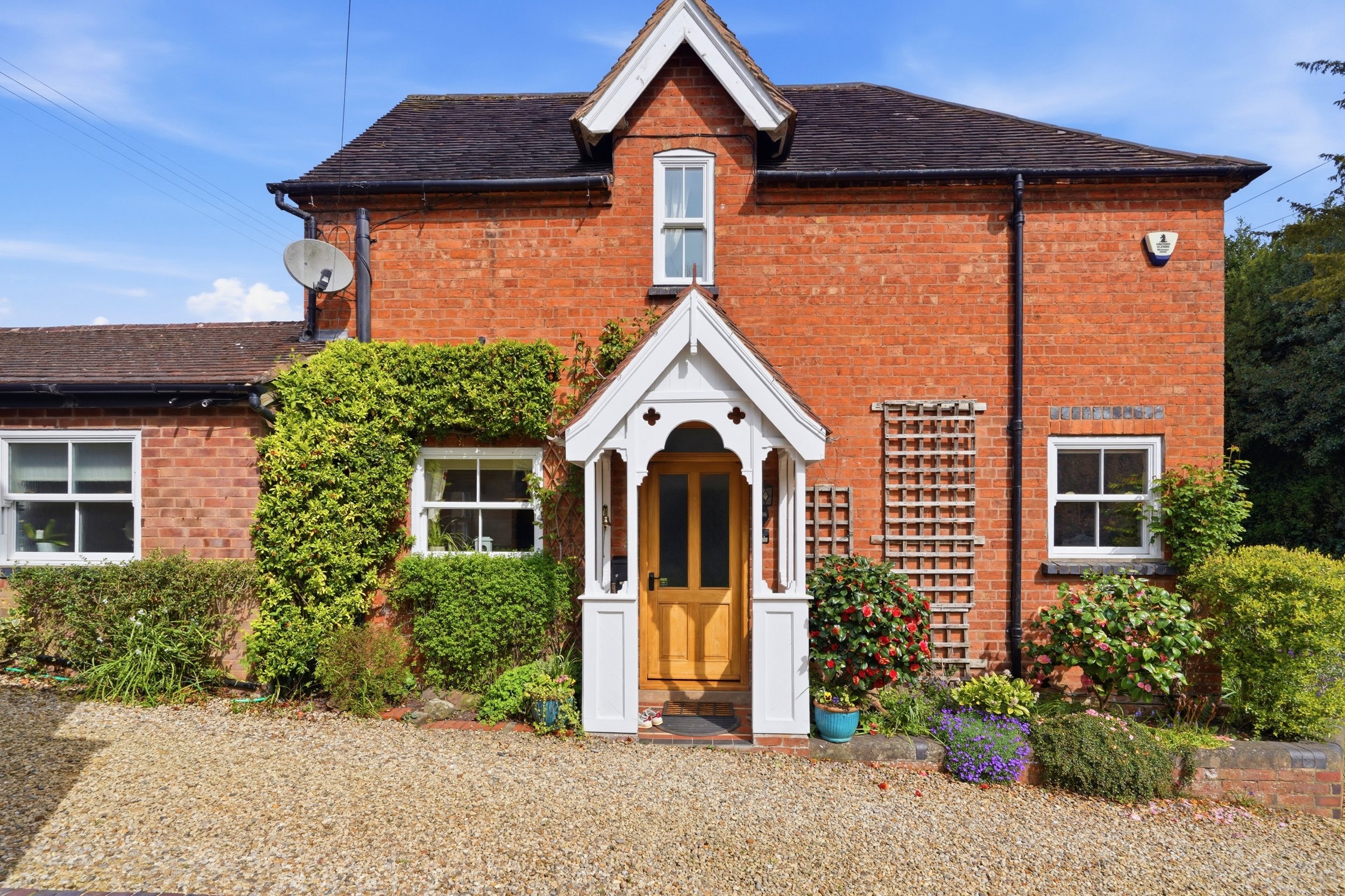 Front view of a traditional brick house with a white decorative porch, surrounded by green shrubs and colorful flowers, under a bright blue sky.