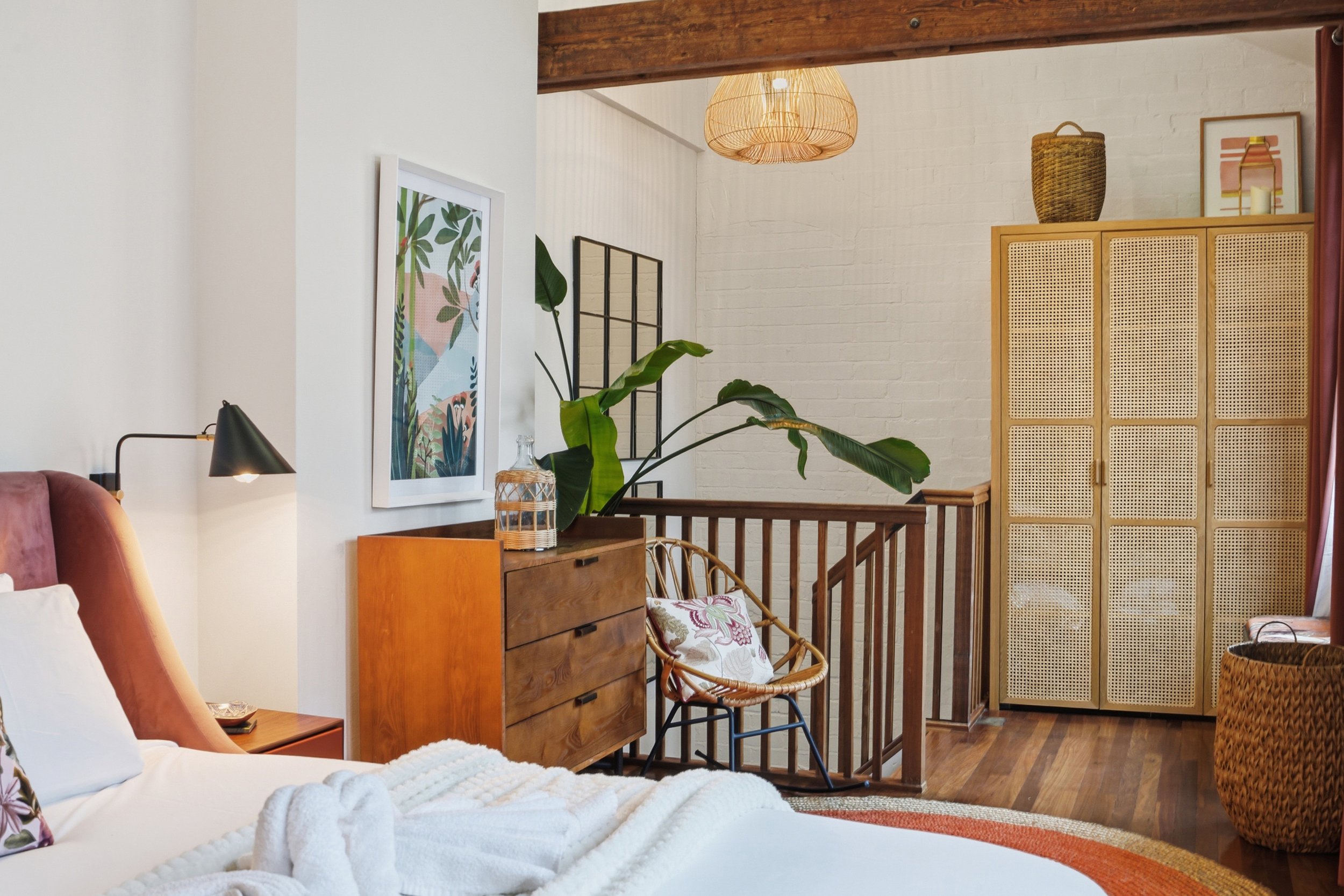 A cozy corner of a living space featuring a bed with white linens, a pink velvet armchair, a wooden dresser with a glass jar on top, a large leafy green plant, framed artwork on the wall, a rattan ceiling light, and a wooden room divider with a woven