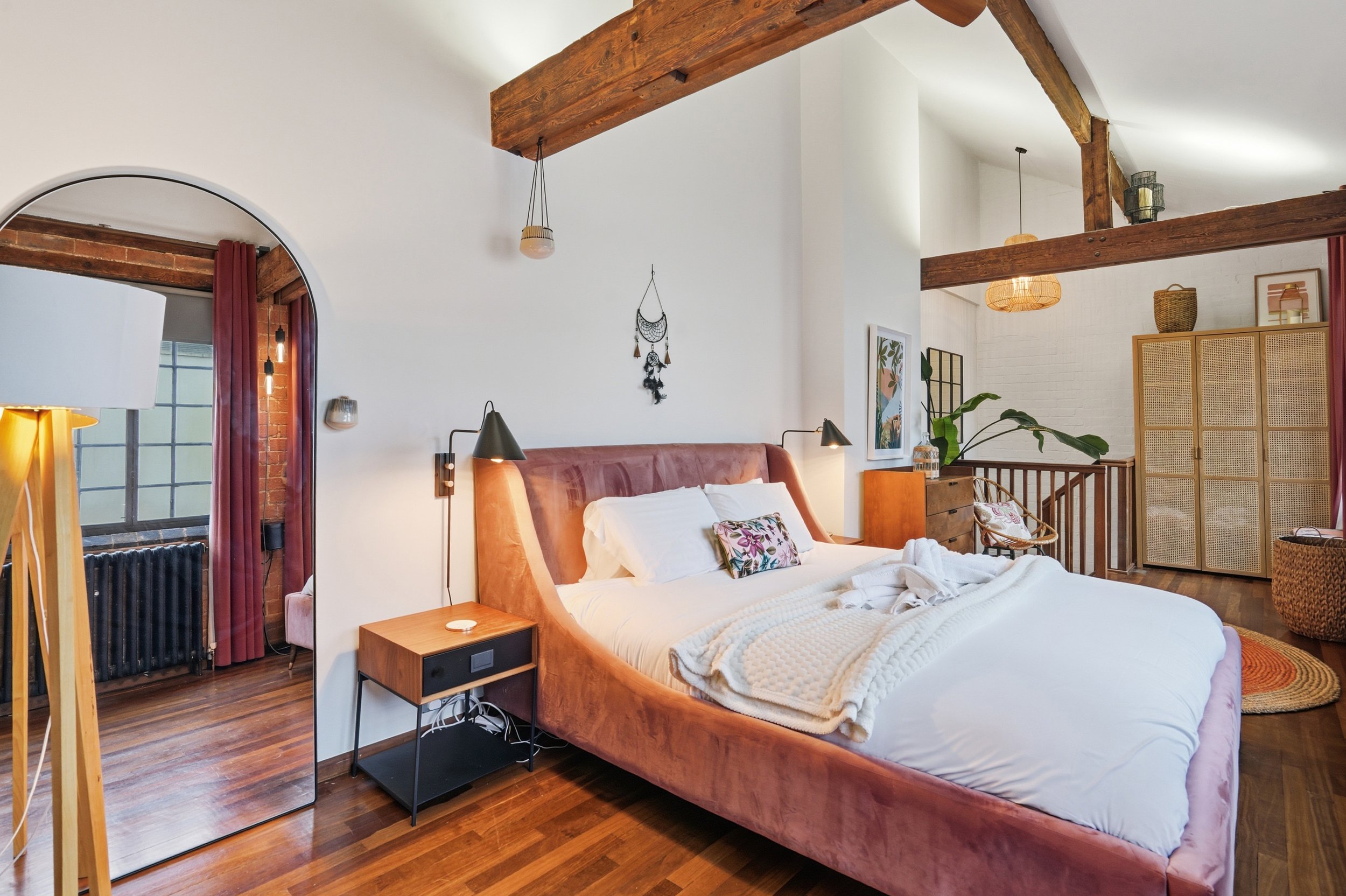 A cozy bedroom with a pink upholstered bed, white bedding, and decorative pillows. Wooden beams ceiling, wall-mounted lamps, framed artwork, plants, and natural decor elements. Hardwood floor with a circular woven rug.