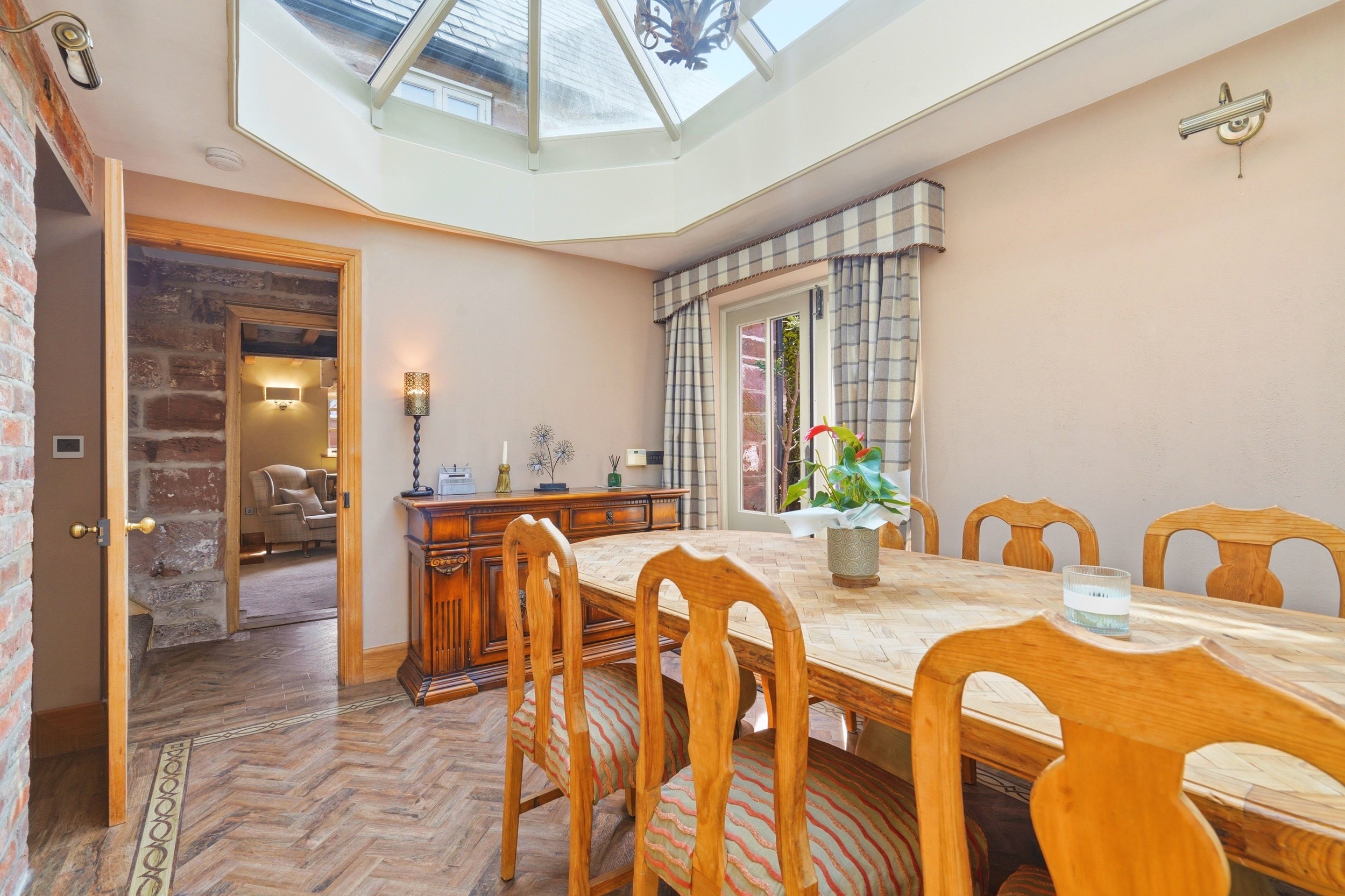Dining room with wooden table and chairs, plants, chandelier, windows with plaid curtains, and a skylight ceiling.