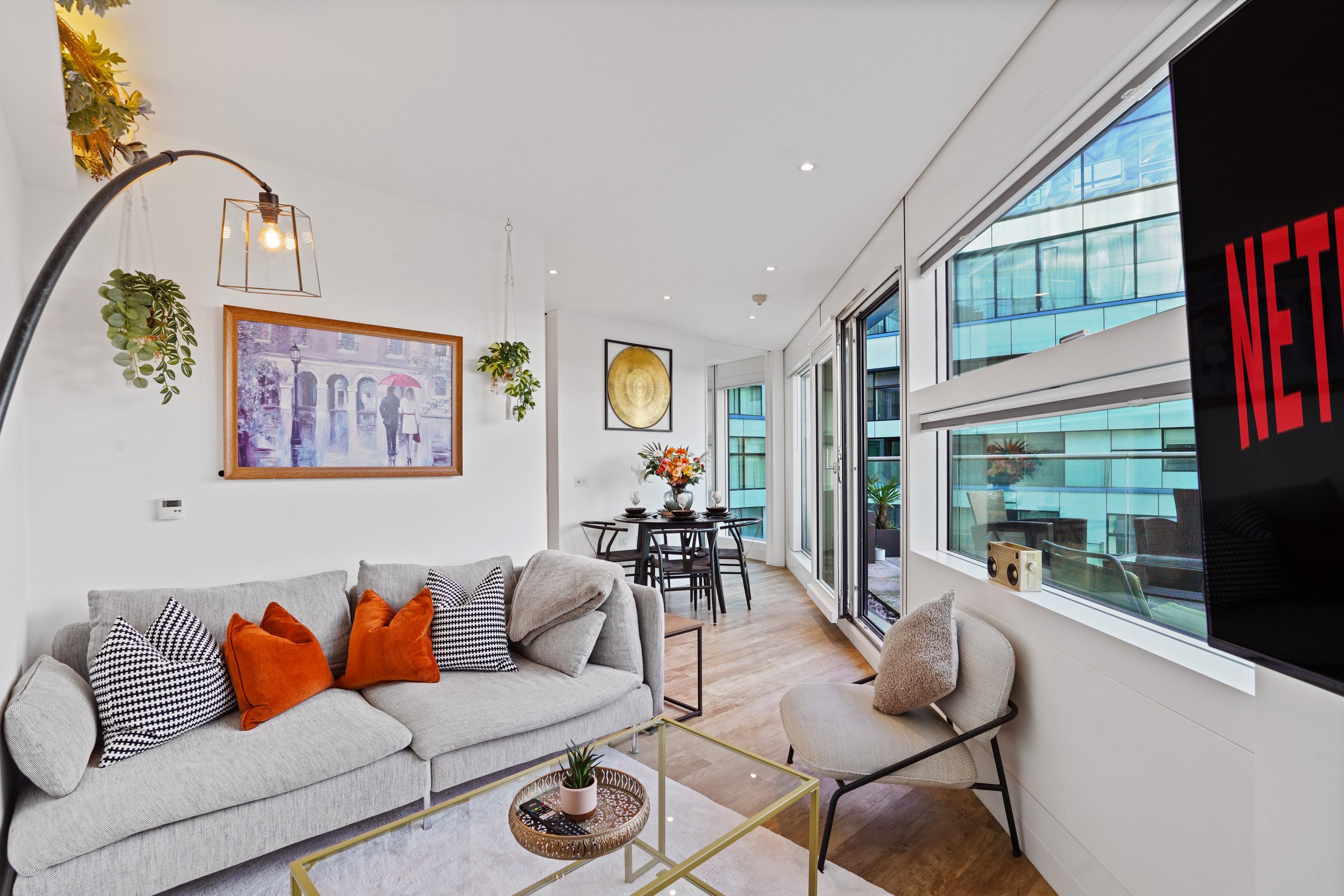 Modern living room with light gray sofa adorned with orange and black-and-white patterned pillows, a coffee table with a plant and remote, and a dining area with a black table and chairs, large windows, wall art, and decorative lighting.