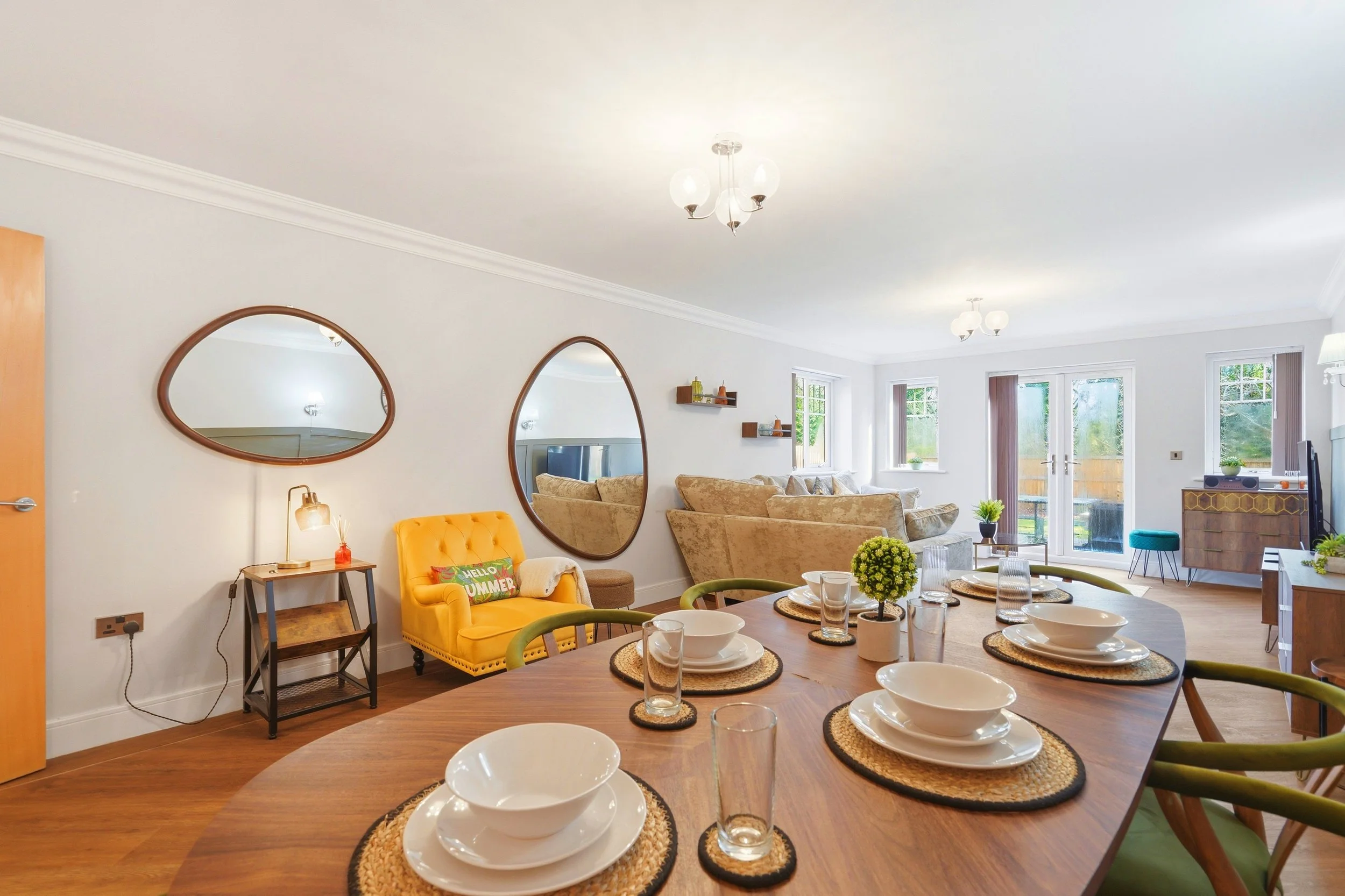 A dining room with a round wooden table set with white bowls, glasses, and doilads. The room has large windows, white walls, and modern lighting fixtures. There is a yellow armchair with a pillow that reads 'Hello Summer,' and a beige sofa in the bac