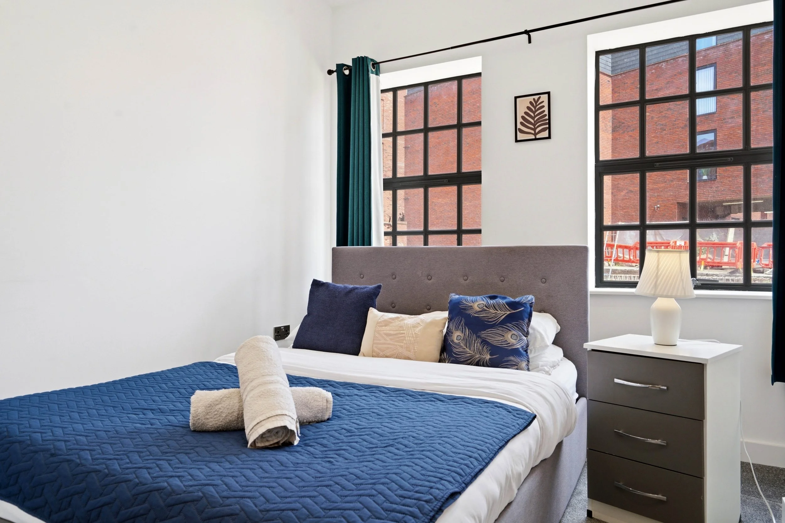 Bedroom with large windows showing red brick building outside; bed with white bedding and blue quilt, decorative pillows, towel rolled at foot of bed; bedside white table with lamp; framed leaf artwork on wall; teal curtain on window.