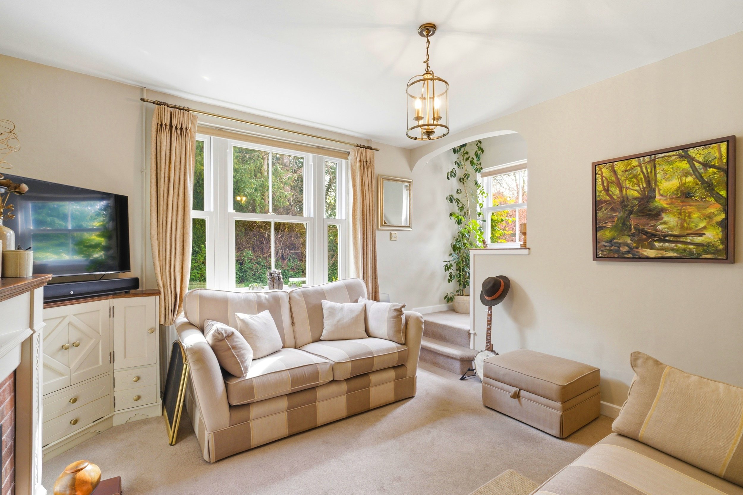 A cozy living room with large windows, a beige striped sofa, a beige armchair, a patterned ottoman, a wall-mounted flat-screen TV, a framed landscape painting, and a tall houseplant near the window.