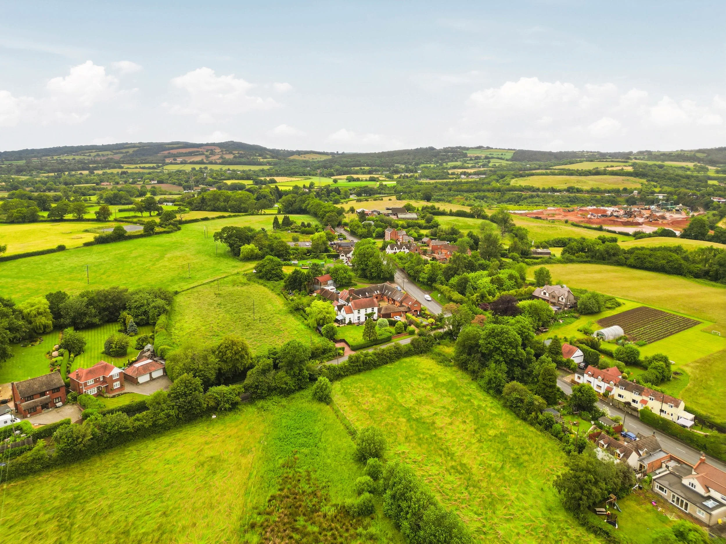 A scenic view of lush green fields, scattered houses, and rolling hills under a partly cloudy sky.