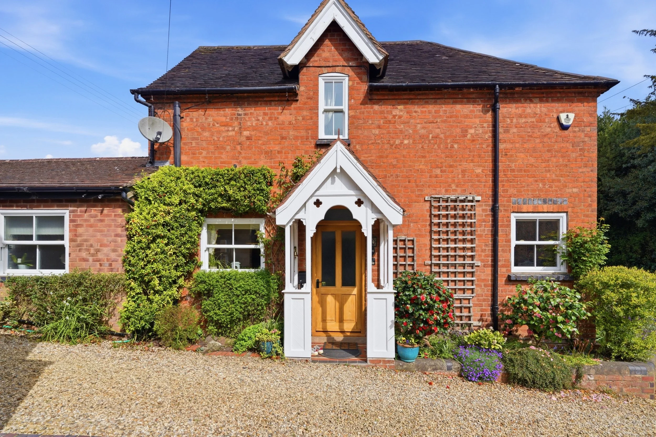 Red brick house with a white front porch and door, surrounded by green plants and flowering bushes, under a blue sky with some clouds.