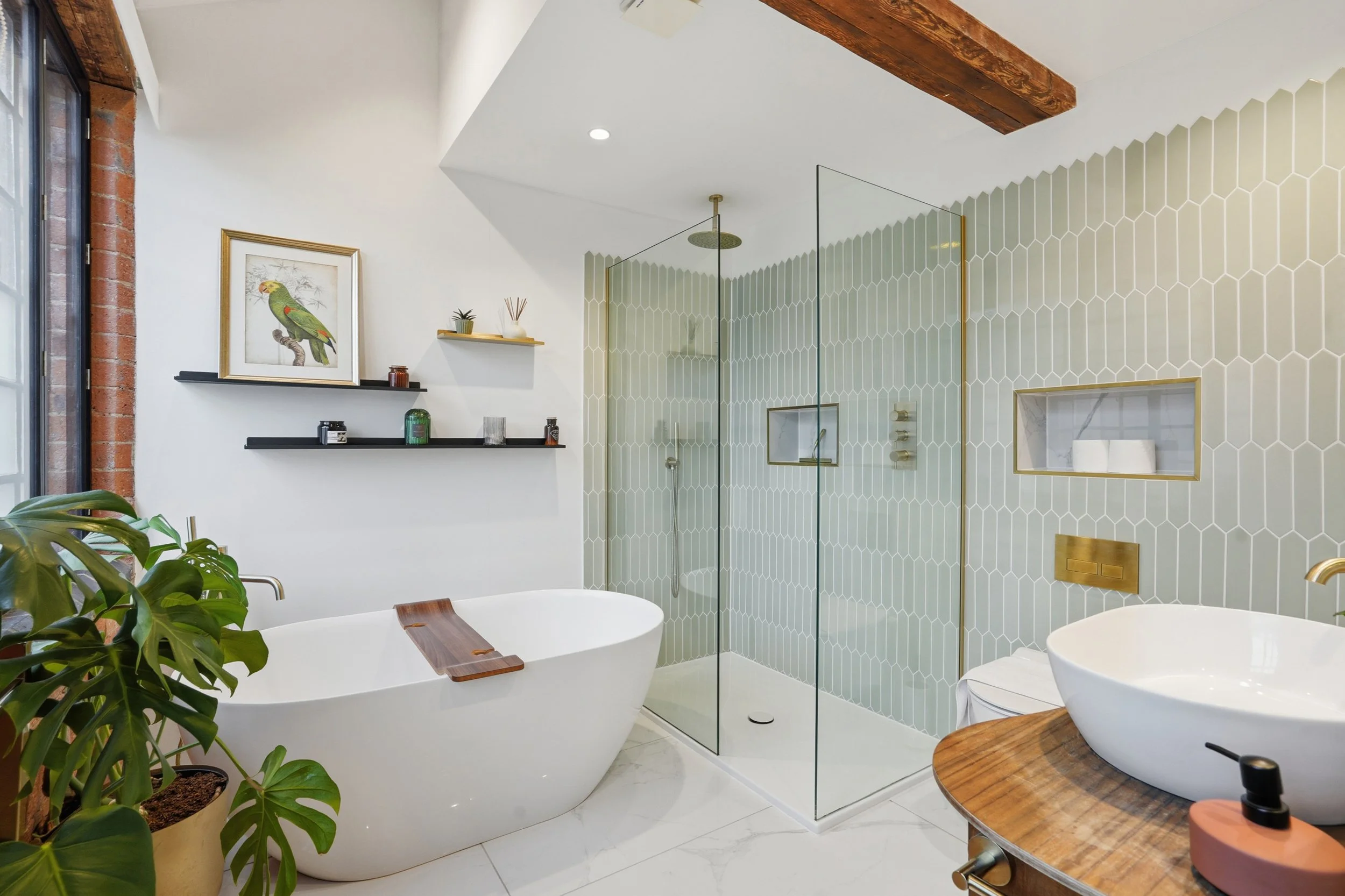 Modern bathroom with a freestanding bathtub, a glass shower enclosure, and greenery including potted plants. Wall shelves hold decor and toiletries, with a framed parrot illustration above. Wooden beams and marble accents add warmth.