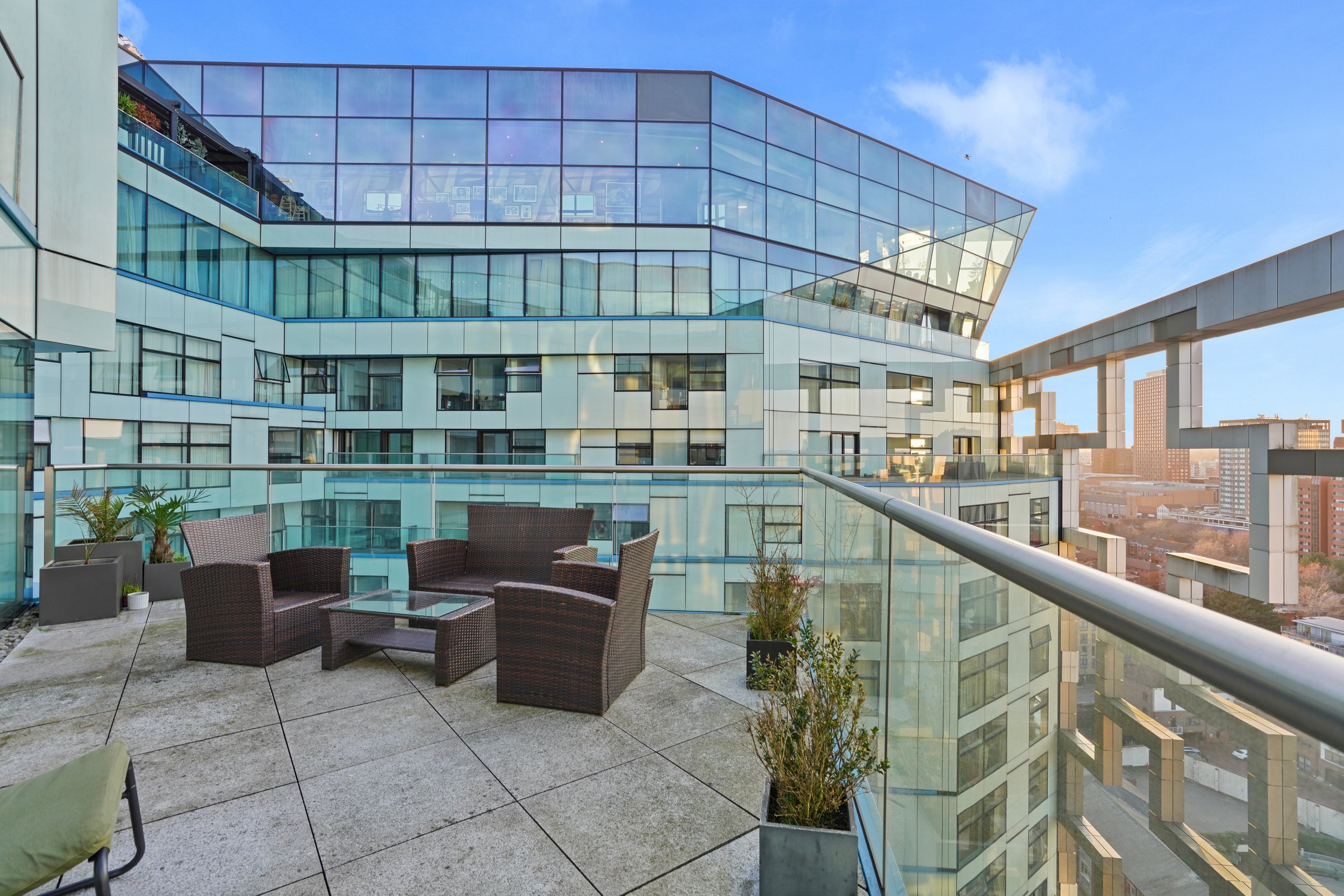 Outdoor balcony with wicker chairs and potted plants, overlooking modern glass building and city skyline under blue sky.